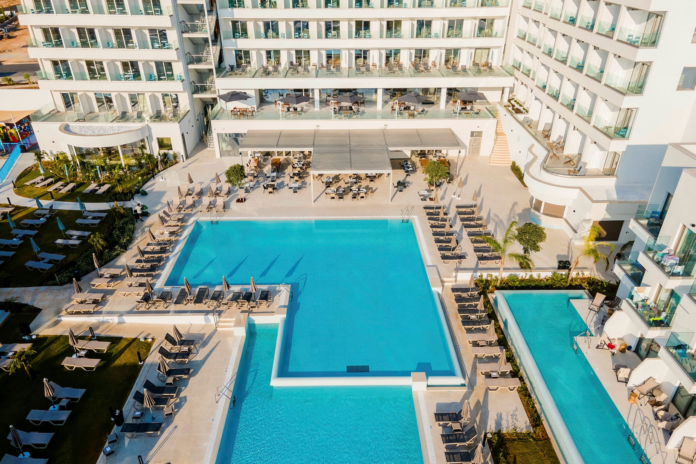Vy av hotellpoolområdet med solstolar och fasaden av en byggnad med balkonger. (AI-genererad text)