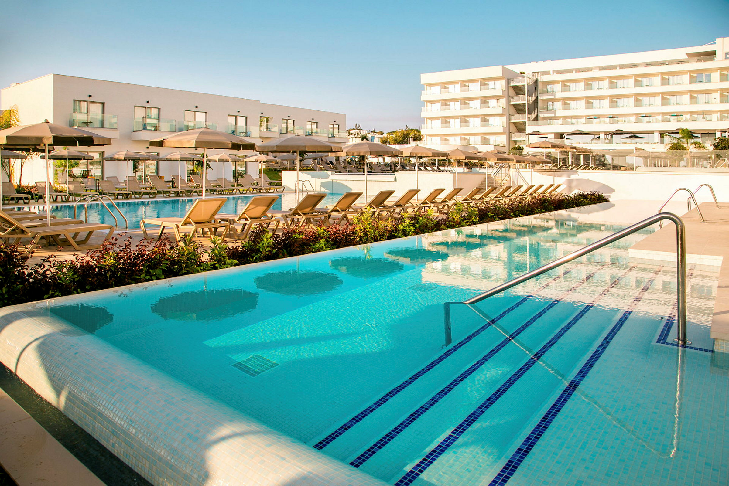 Poolområde med solstolar och parasoller framför moderna hotellbyggnader. (AI-genererad text)