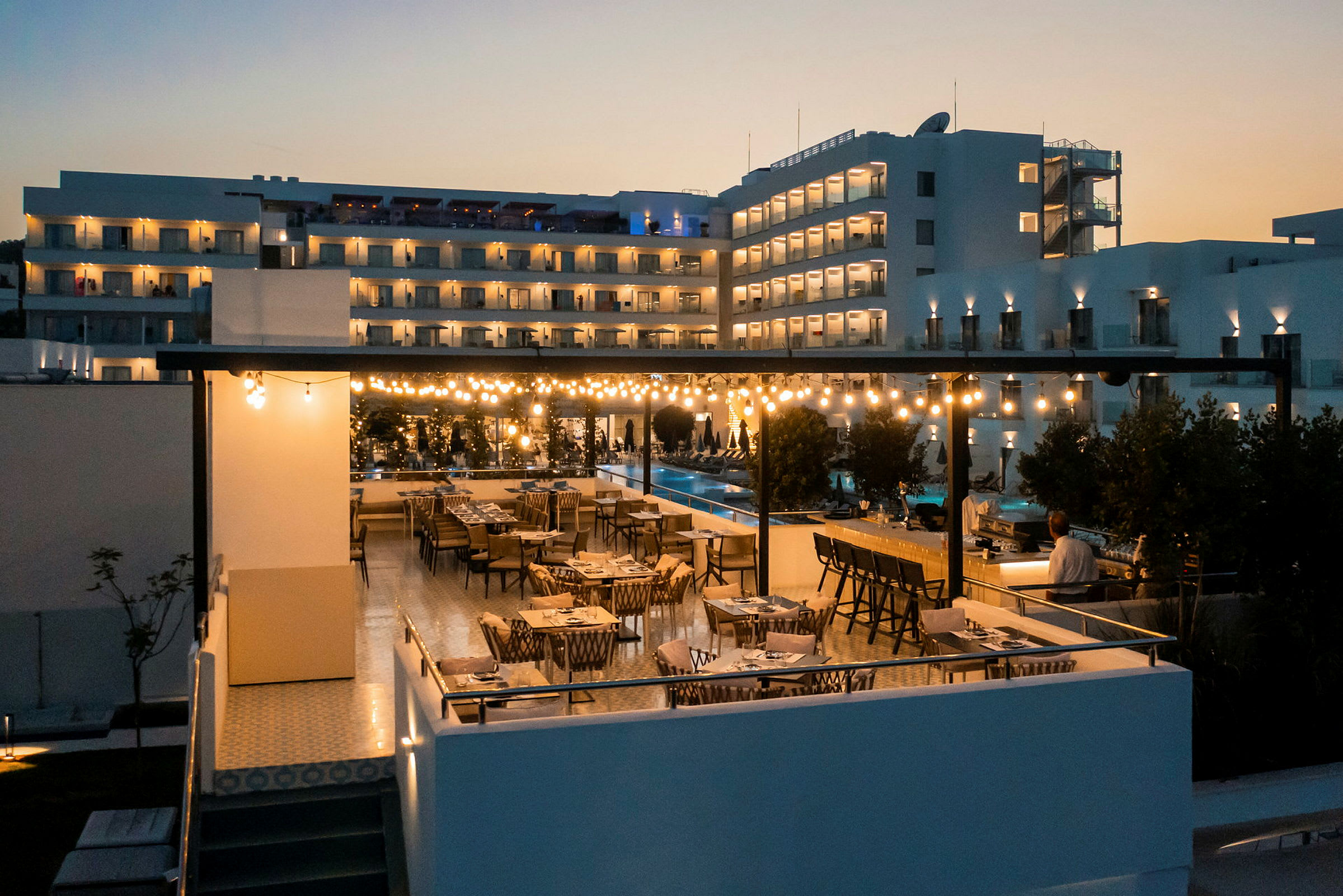 En restaurangterrass med bord och stolar på kvällen, upplyst av ljusslingor, med en modern byggnad i bakgrunden. (AI-genererad text)