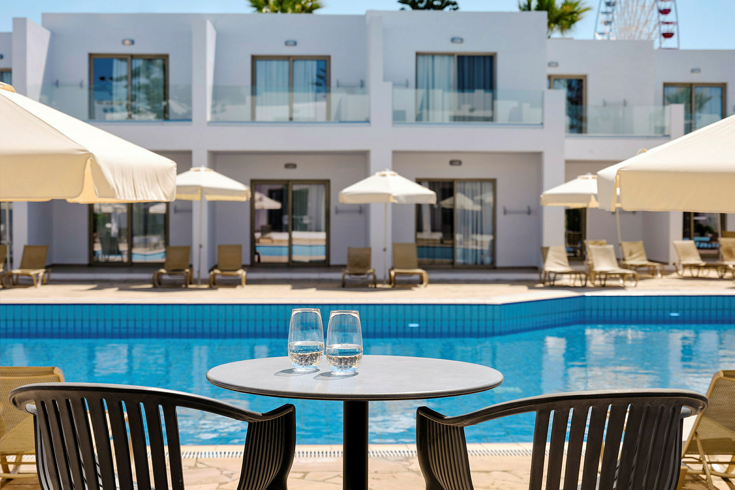 Utsikt över en pool med solstolar och parasoller, samt ett bord med två glas vatten i förgrunden. (AI-genererad text)