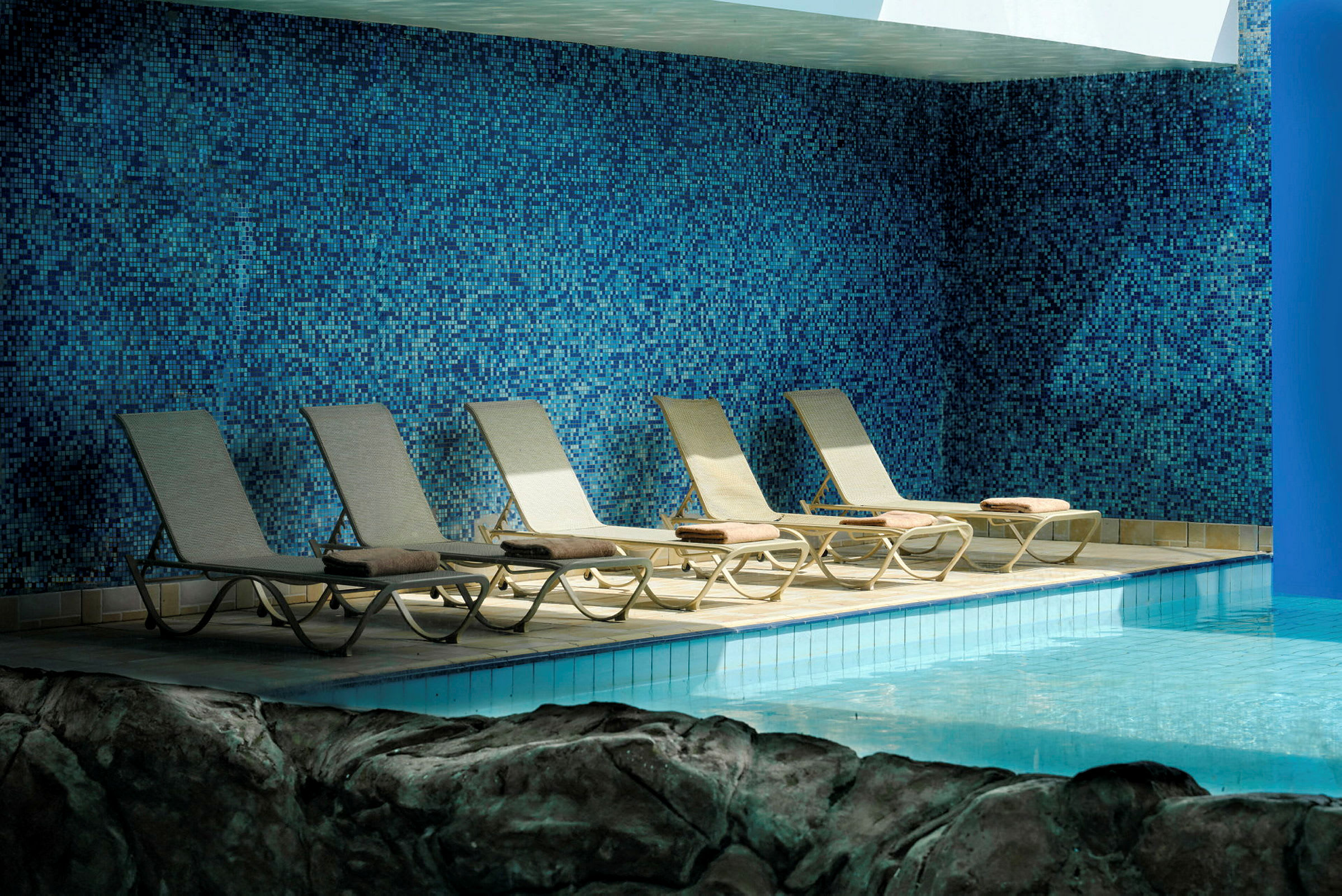 Solstolar vid kanten av en pool, med en blå mosaikvägg bakom. (AI-genererad text)