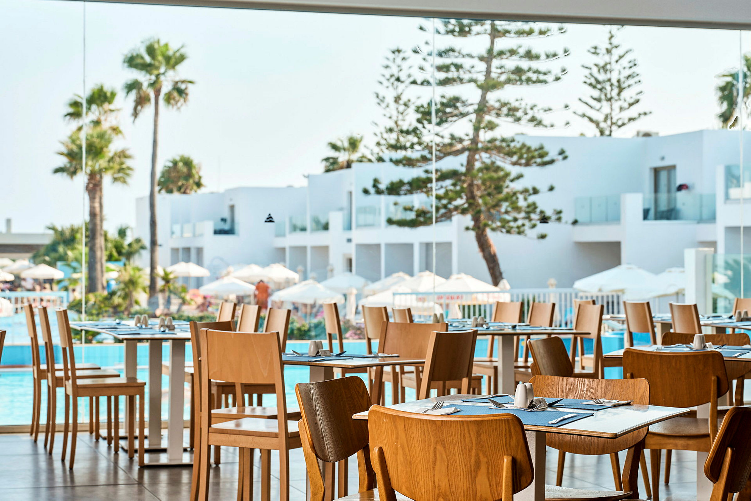 Restaurang med trästolar och dukade bord, utsikt mot vit arkitektur, pool och palmer. (AI-genererad text)
