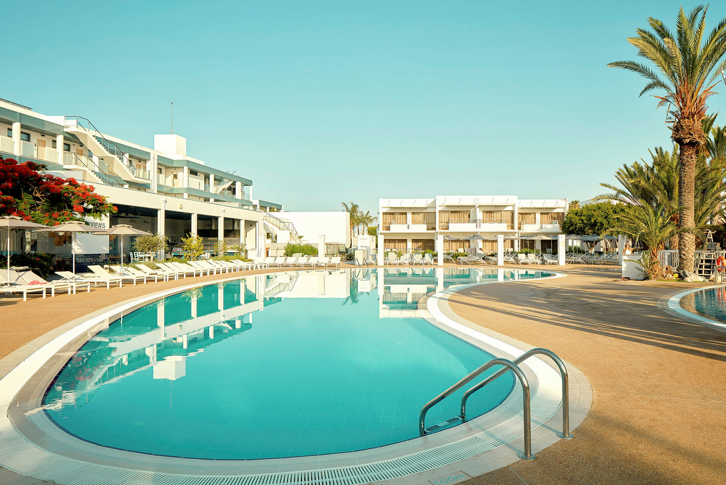 Ett hotellområde med en stor pool och omgivande solstolar. I bakgrunden syns hotellbyggnader och palmträd. (AI-genererad text)
