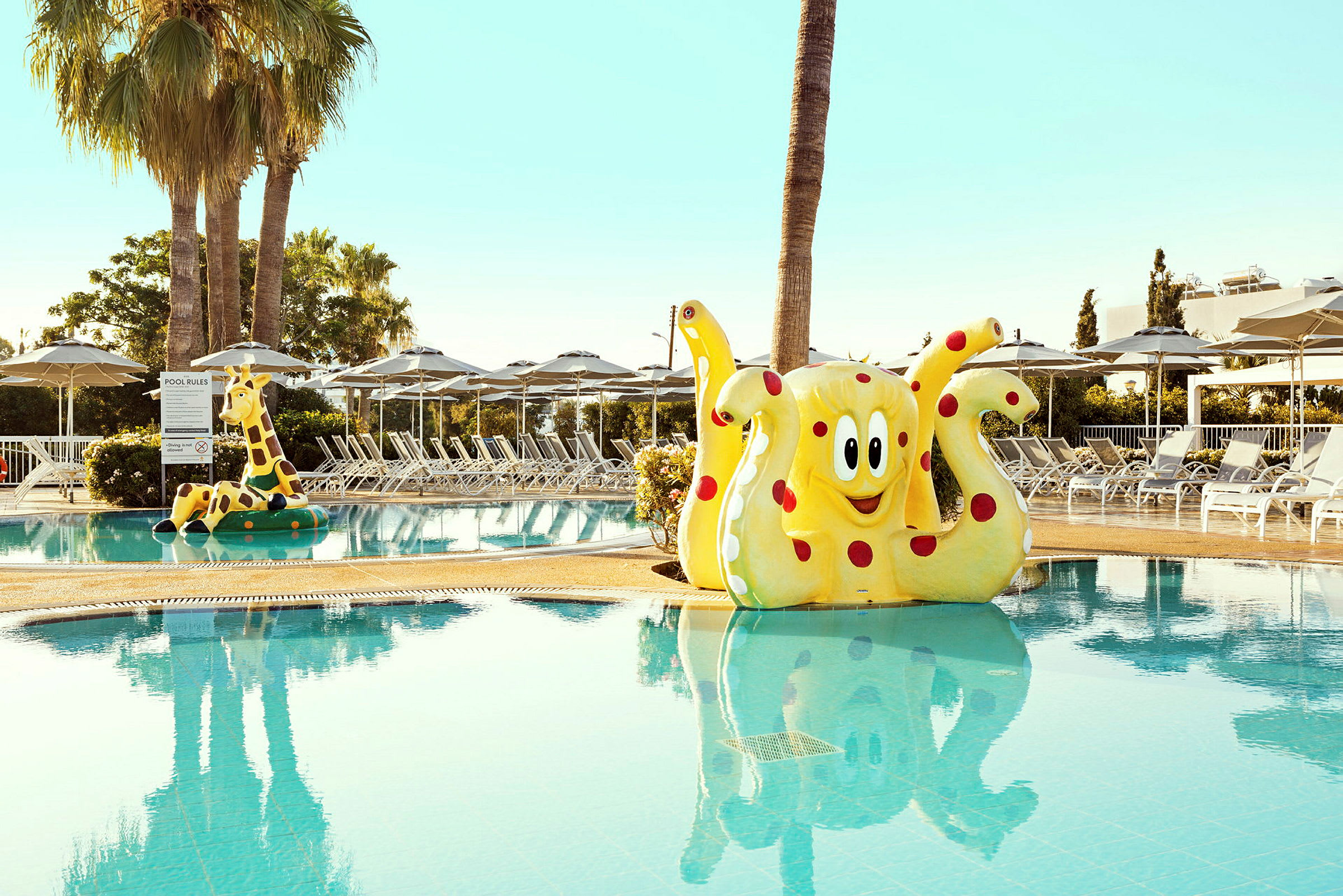 En pool med en gul, rödprickig bläckfiskfigur samt en giraffrutschkana och solstolar i bakgrunden. (AI-genererad text)
