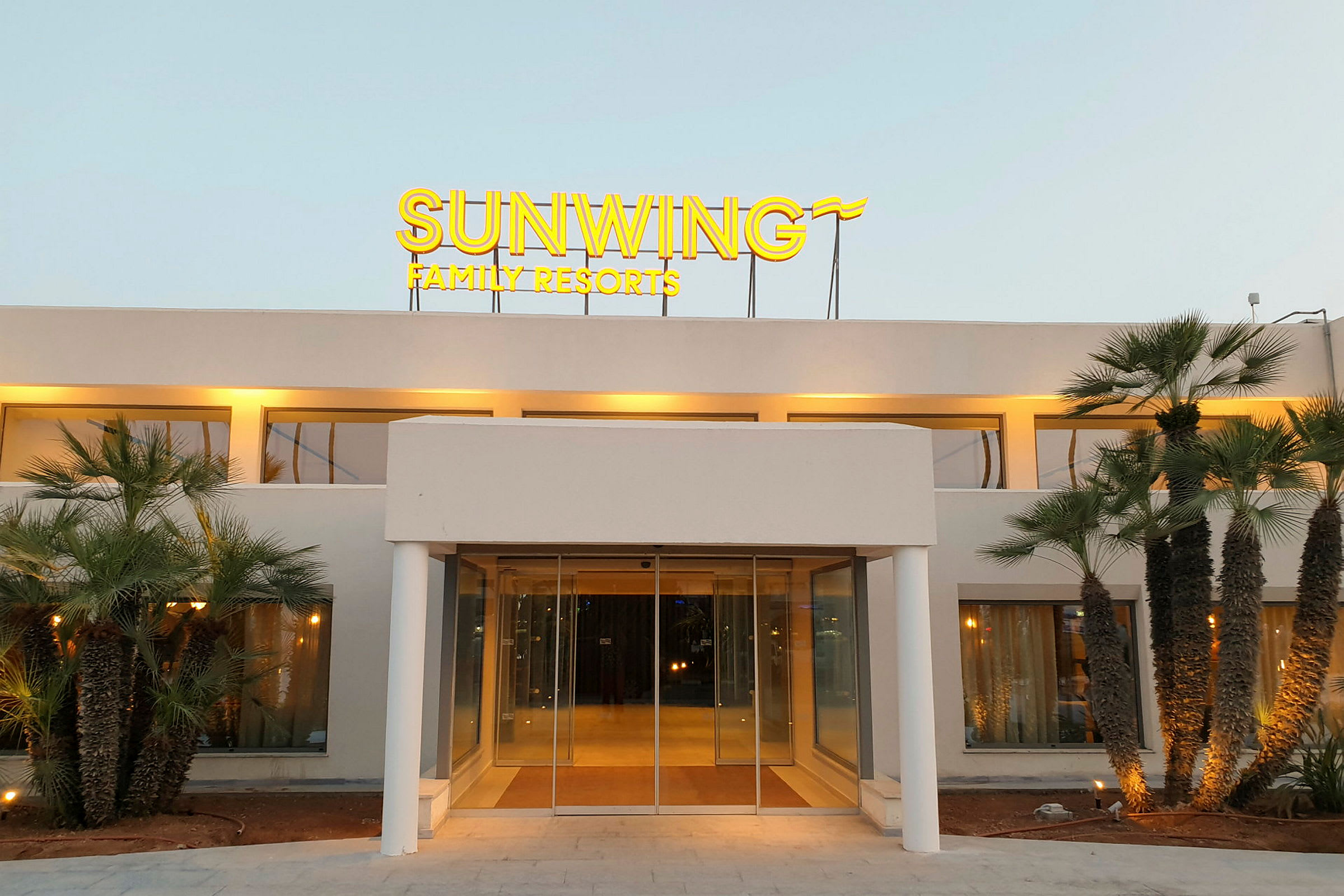 Framsidan av en byggnad med en upplyst skylt som lyder "Sunwing Family Resorts" och omgiven av palmer. (AI-genererad text)
