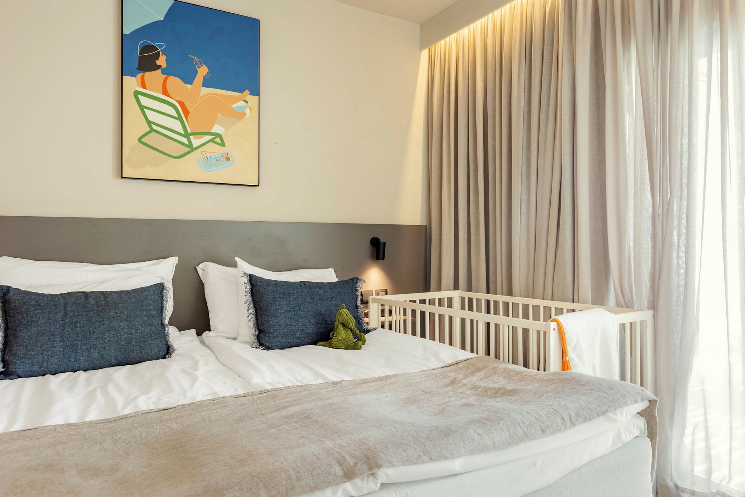 Ett hotellrum med en dubbelsäng, prydd med blå kuddar och en illustration på väggen ovanför. En babysäng syns bredvid fönstret med tunna gardiner. (AI-genererad text)