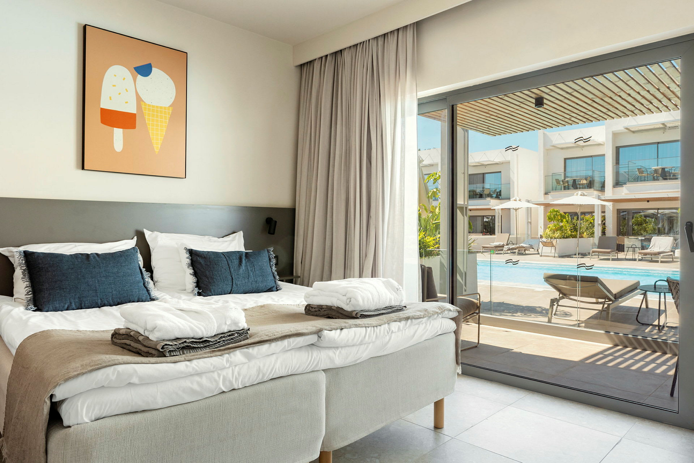 En hotellrum med en dubbelsäng, ljust grå sänggavlar och blå kuddar. En tavla med glassmotiv hänger på väggen. Genom glasdörren syns en utomhuspool med solstolar och parasoller. (AI-genererad text)