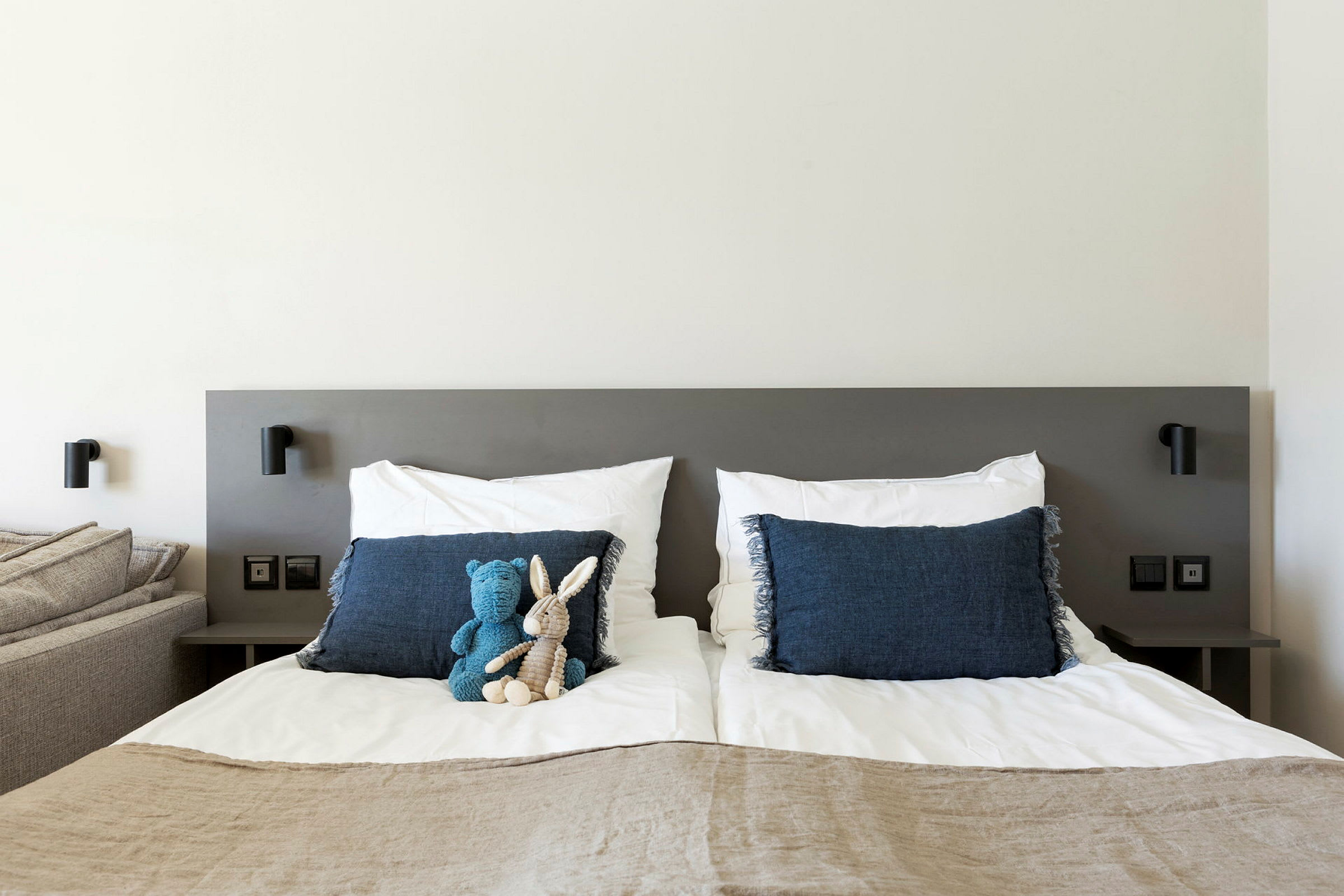 Bilden visar en modern sovrumsmiljö med en dubbelsäng klädd i vita sängkläder och blåa kuddar. På sängen ligger två mjukdjur, en blå och en beige. Vägglampor är monterade vid sänggaveln. (AI-genererad text)