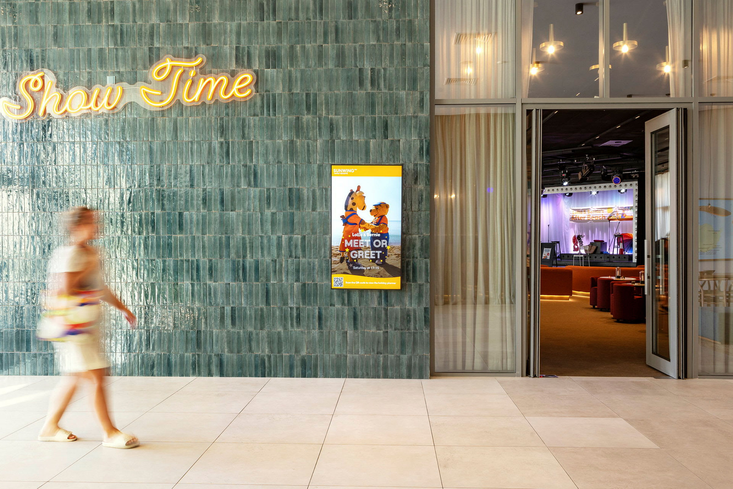 En person går förbi en vägg med texten "Show Time" och en affisch för ett barnprogram. I bakgrunden syns en scen med publikstolar. (AI-genererad text)