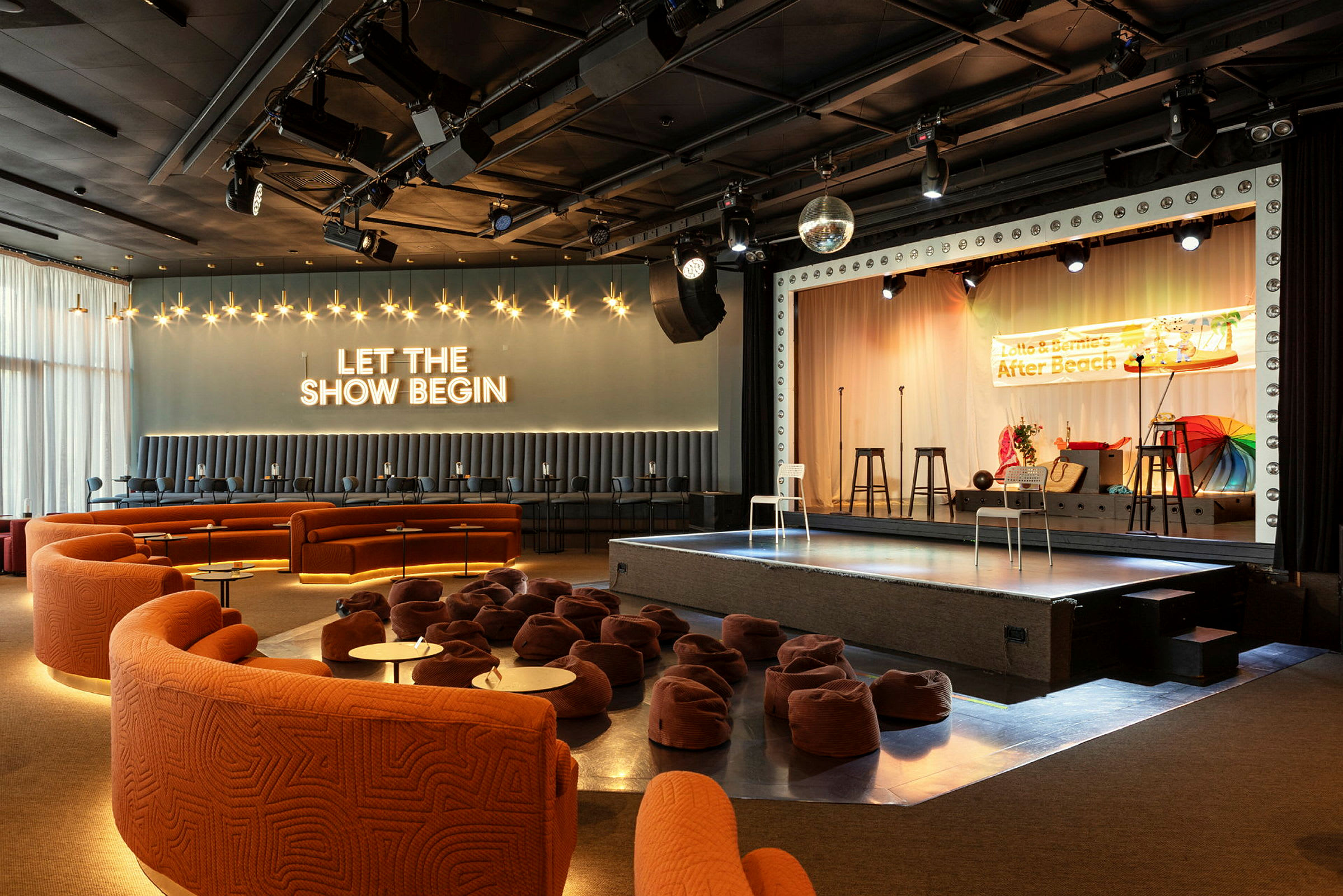 Ett rum med scen, sittplatser och dekorationer. Texten 'LET THE SHOW BEGIN' lyser på väggen. (AI-genererad text)