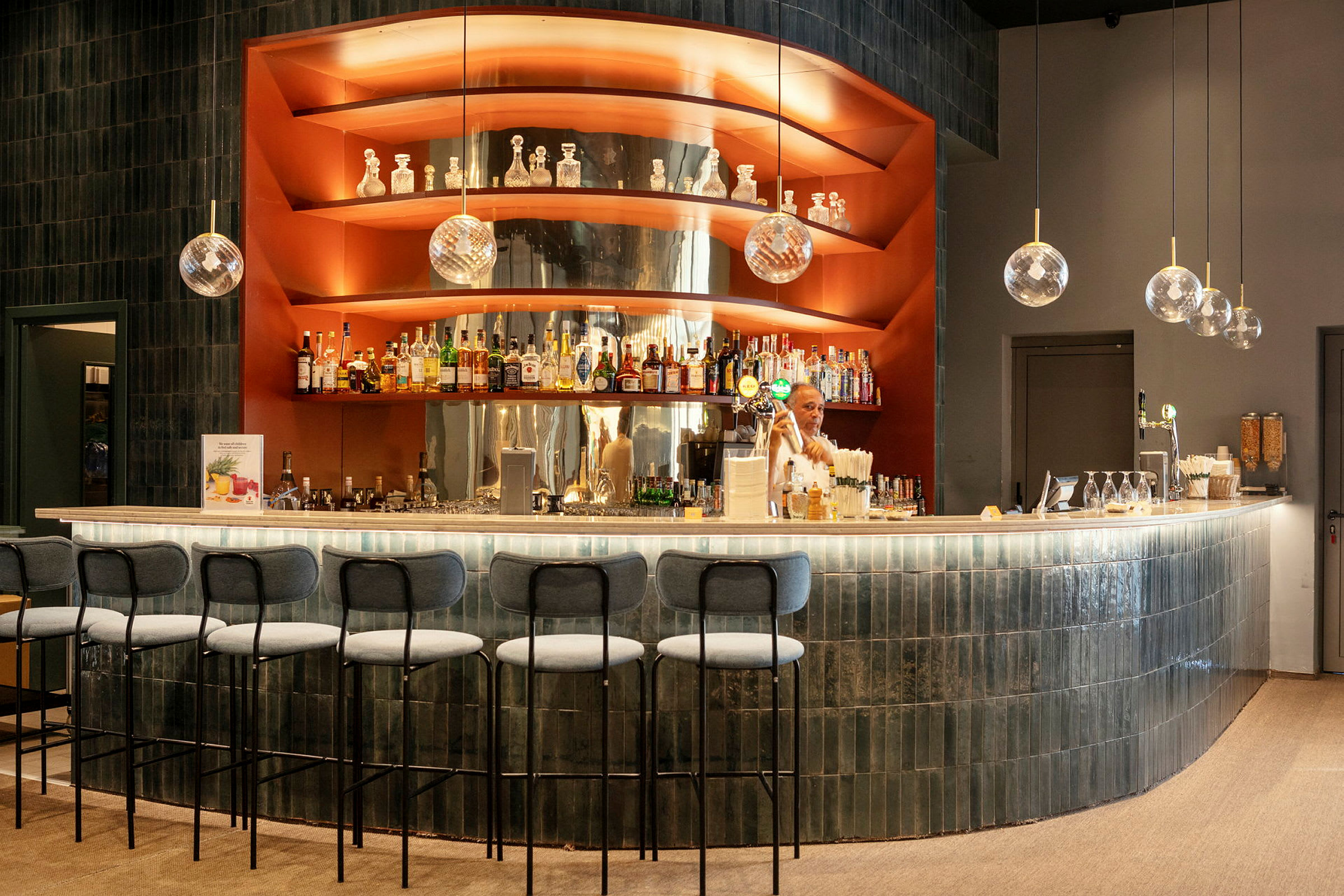 En modern bar med bänkskiva i ljus sten och hyllor med spritflaskor, dekorationer och hängande glaskupor. (AI-genererad text)