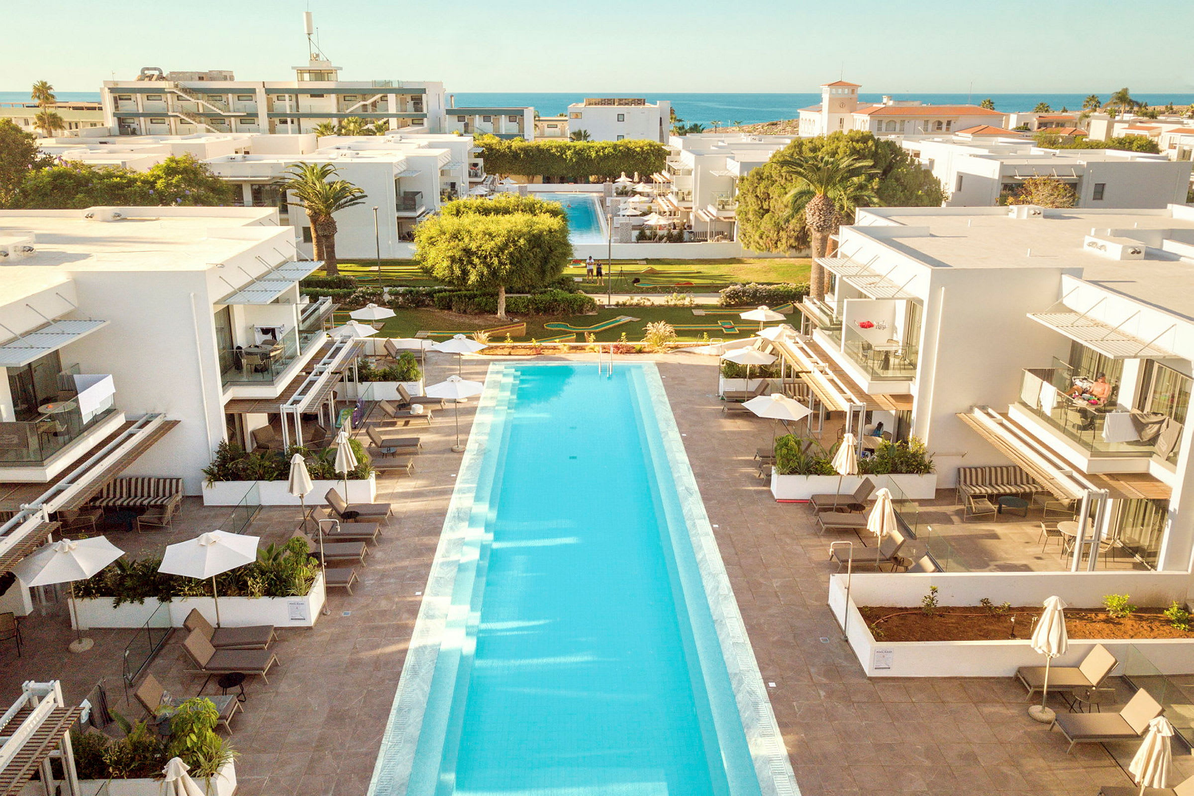 Ett hotellområde med en lång swimmingpool mellan två vita byggnader. I bakgrunden syns grönskande gräsmattor och havet. (AI-genererad text)
