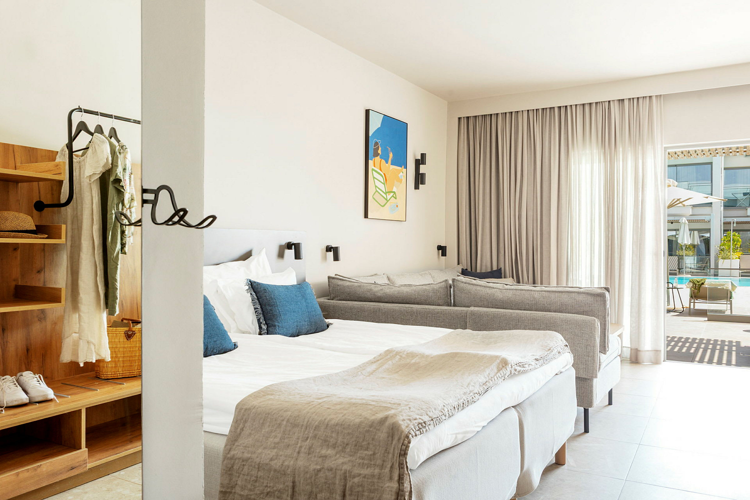 Ett hotellrum med en dubbelsäng med vita lakan och blå dekorativa kuddar, en grå soffa samt ett öppet garderobsutrymme med kläder hängande på galgar. Ett konstverk hänger på väggen och en glasdörr leder ut till ett poolområde. (AI-genererad text)