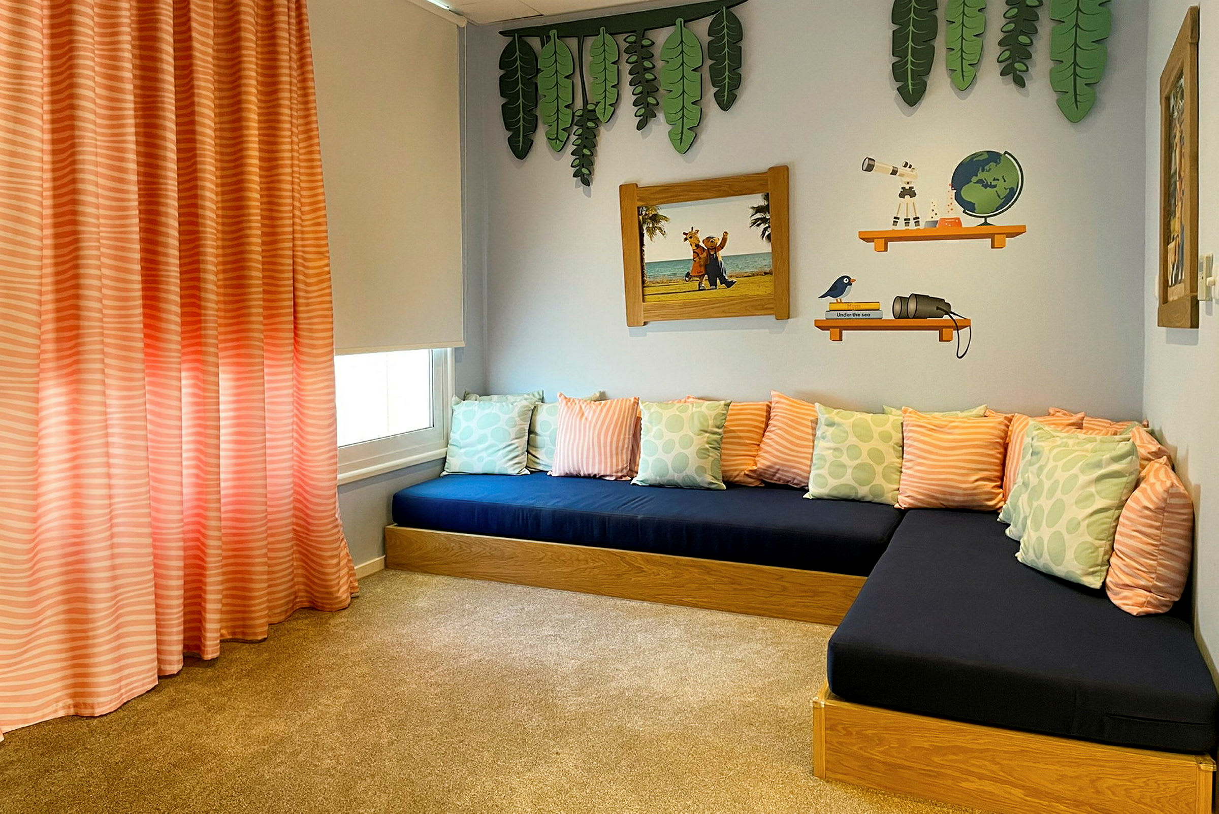 Ett rum med blå sittdynor och flera kuddar i rosa och gröna mönster. Dekorationer på väggarna inkluderar bladväggskonst, ett illustrerat hyllmotiv och en inramad bild med strandmotiv. (AI-genererad text)