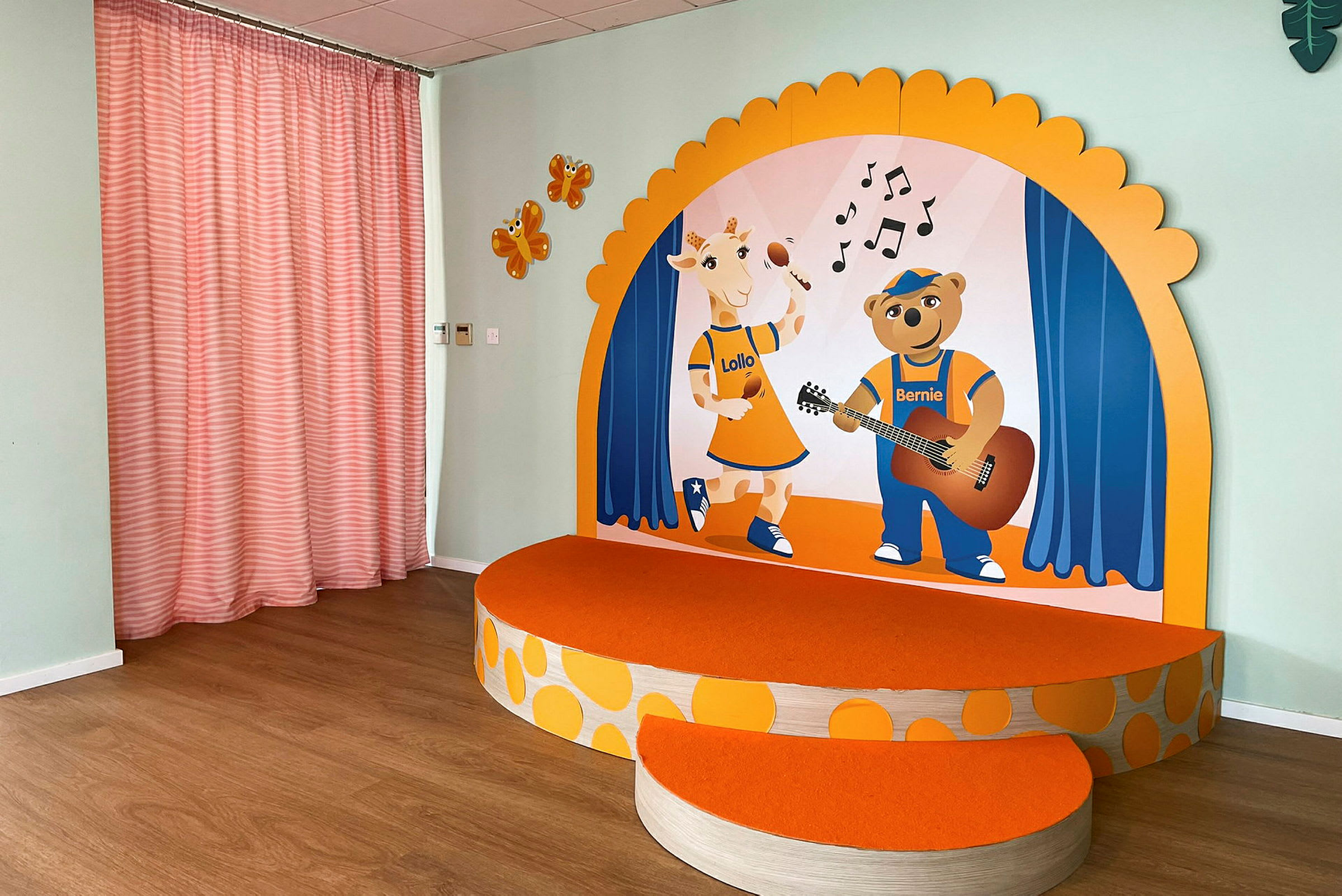 Ett litet upphöjt podium med orange yta och illustrationer av en giraff och en björn på en bakgrund med musiktema. På sidoväggen finns orange figurer och rosa gardiner. (AI-genererad text)
