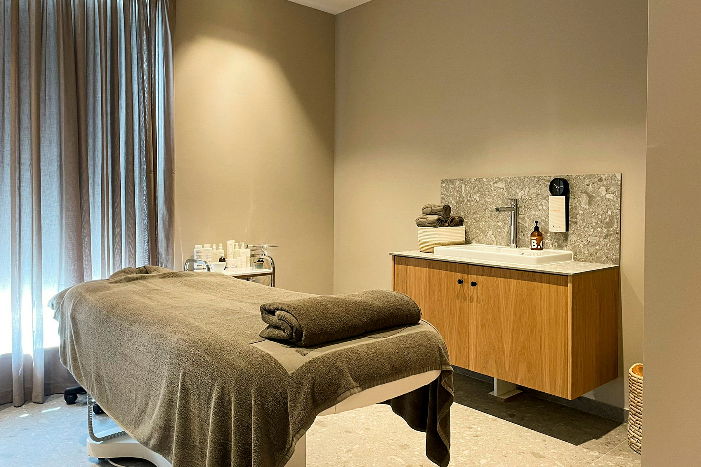 Ett rum med massagebänk klädd med en brun handduk framför ett handfat med träskåp och grå stenbänkskiva. (AI-genererad text)