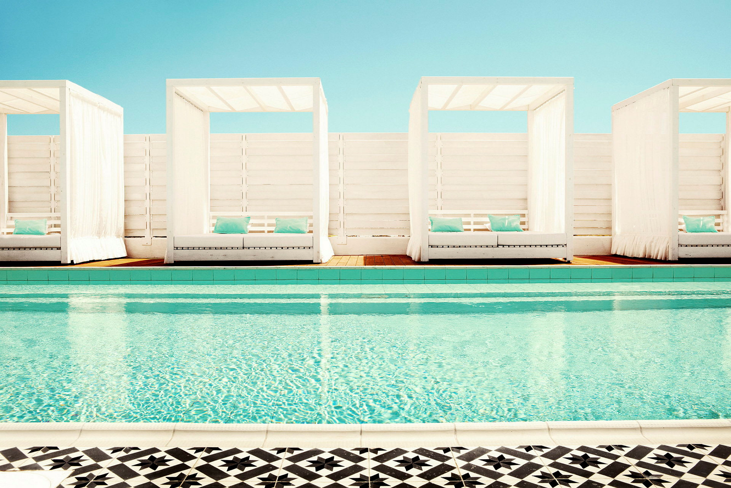 En pool med klart vatten och tre cabanas med vita tyger och blå kuddar i bakgrunden. (AI-genererad text)