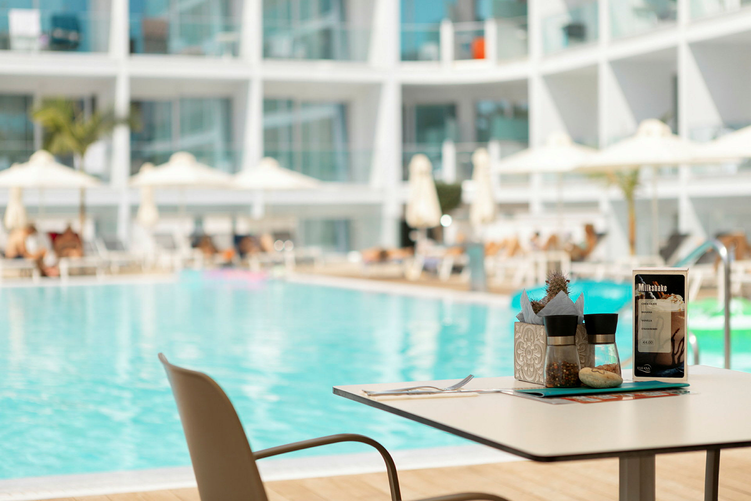 Ett bord med kryddbehållare och meny bredvid en pool med solstolar och parasoller. (AI-genererad text)