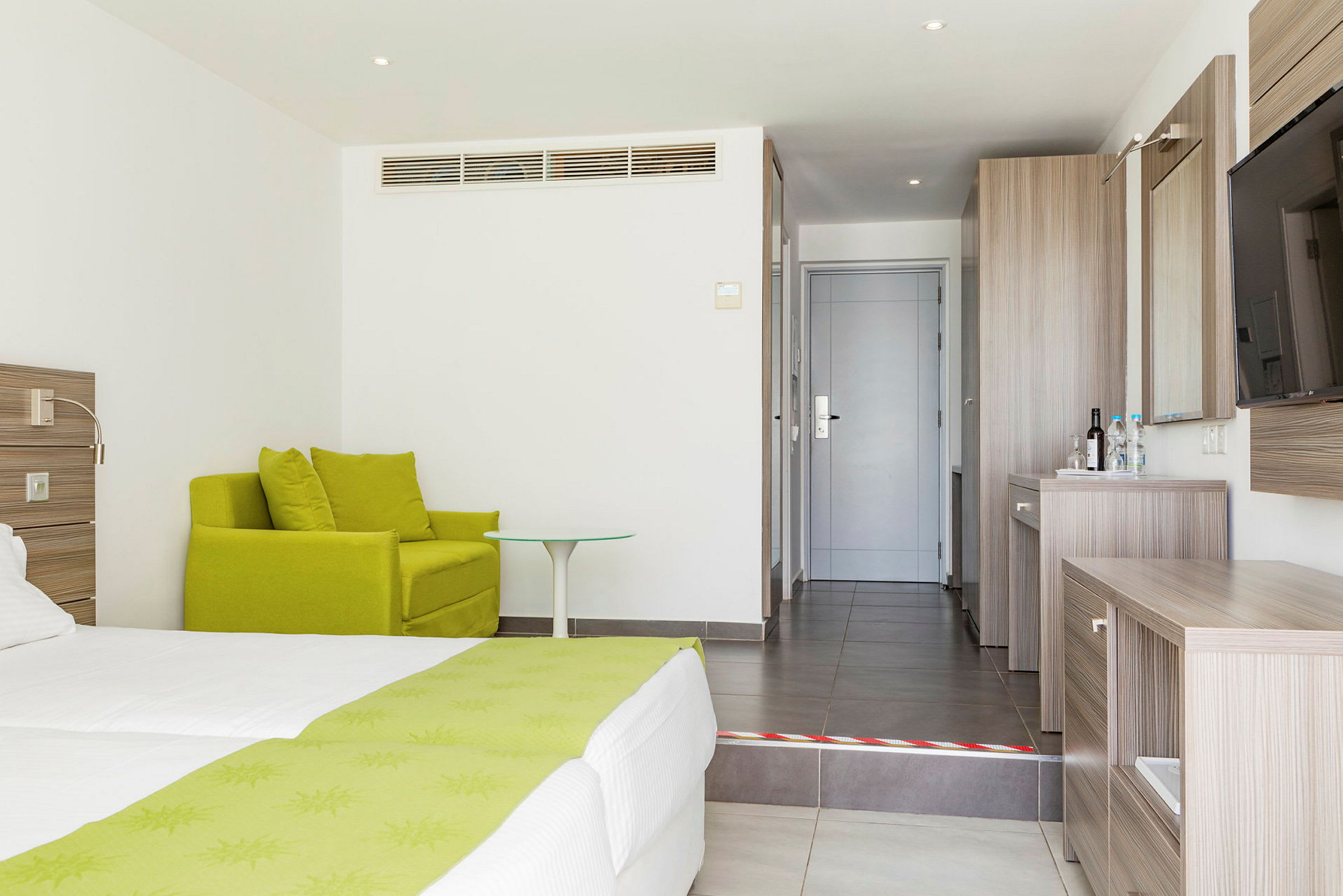Hotellrum med grön fåtölj, säng med gröna detaljer och skrivbord i trä vid ingången. (AI-genererad text)
