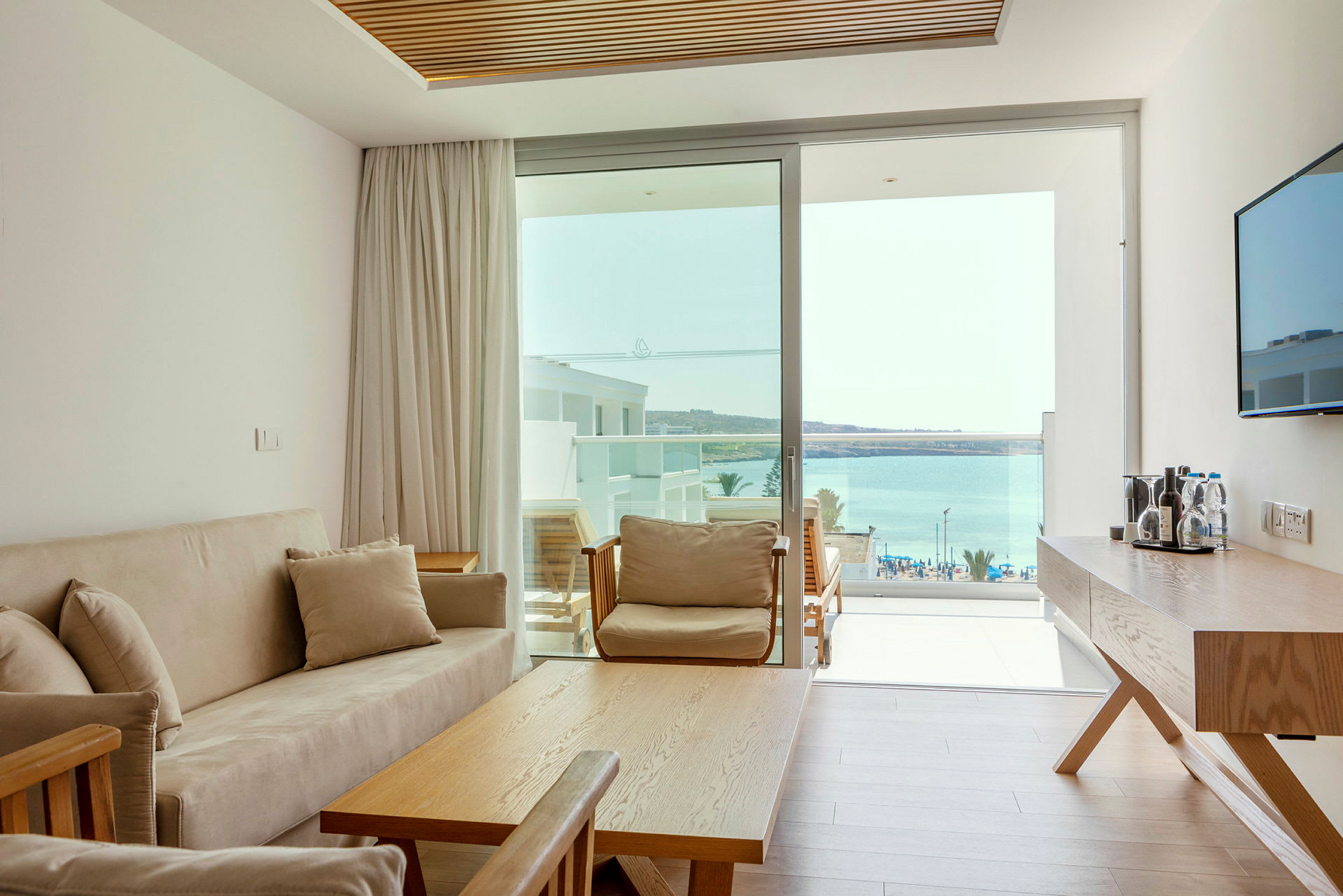 Ett ljust hotellrum med en soffa, bord och utsikt över havet genom glasdörrar. (AI-genererad text)