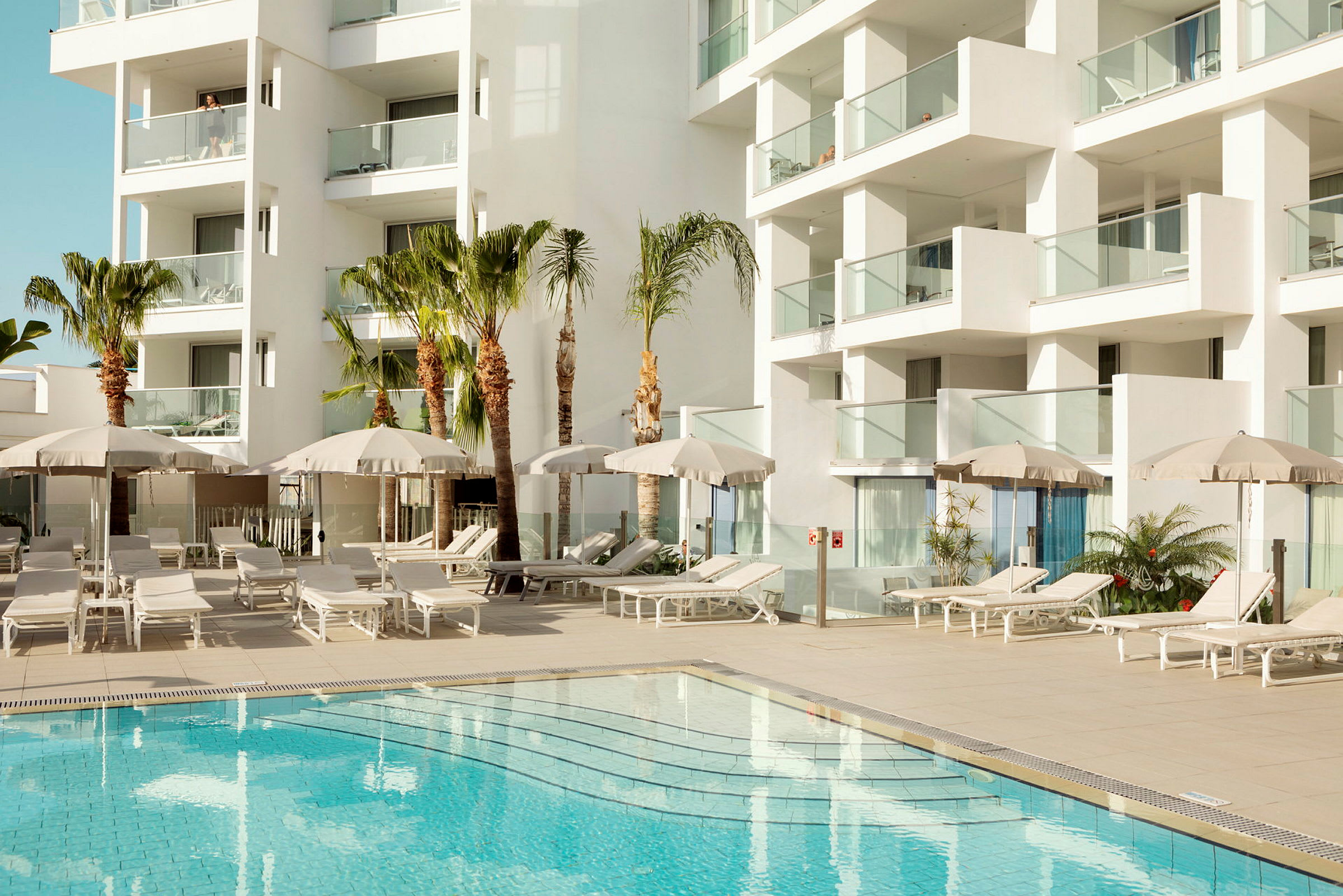 Hotellbyggnad med balkonger samt poolområde med solsängar och parasoll. (AI-genererad text)