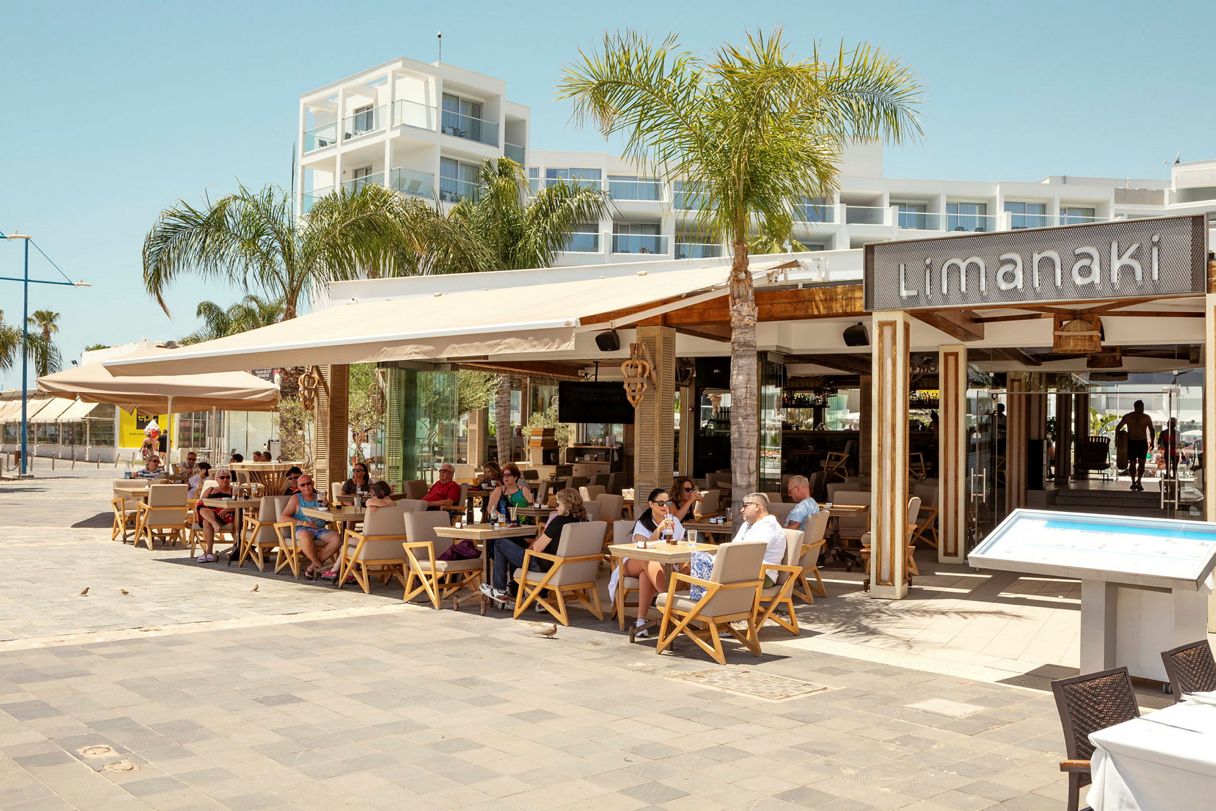Restaurang Limanaki med sittande gäster, omgiven av palmer och modern arkitektur. (AI-genererad text)