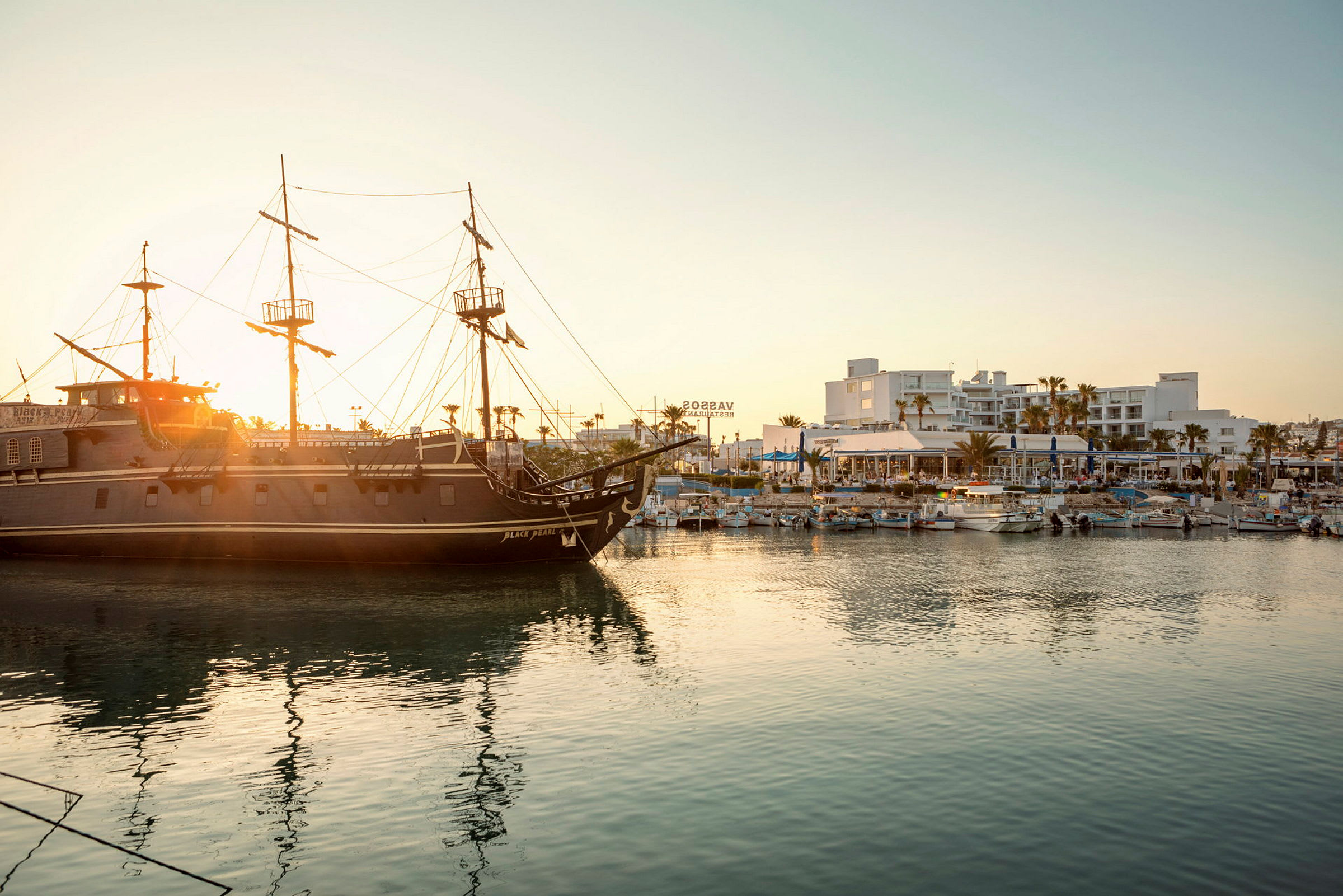 En piratskepp ligger i en hamn med byggnader och båtar i bakgrunden vid solnedgång. (AI-genererad text)