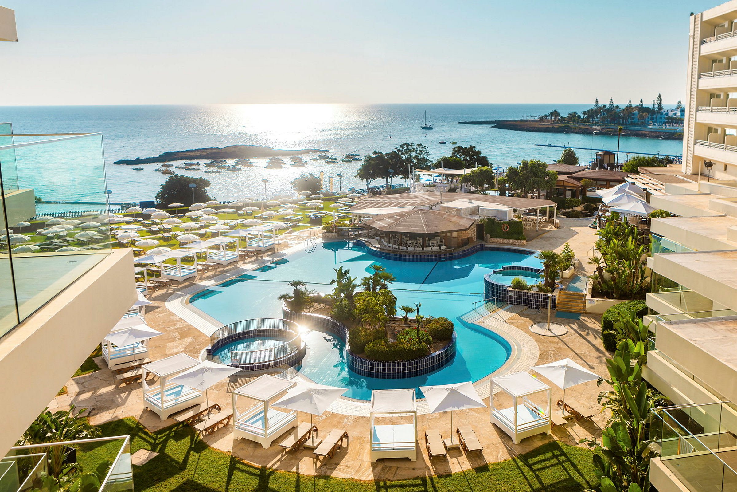 Bilden visar en utomhuspool vid ett hotellkomplex nära havet, omgivet av solstolar, parasoller och grönska. (AI-genererad text)