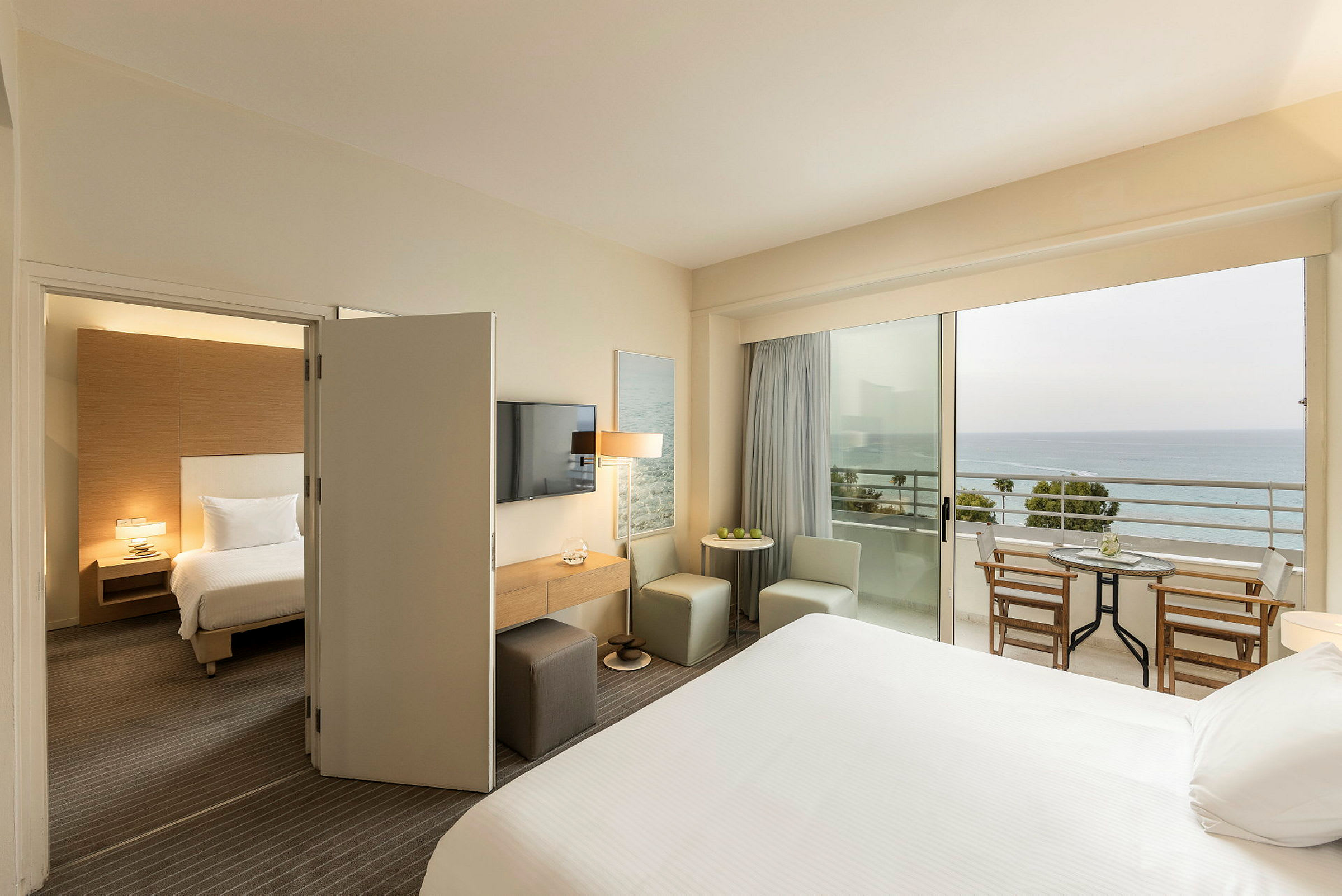Ett hotellrum med en säng, sittplatser och ett bord, och en balkong med utsikt över havet. (AI-genererad text)