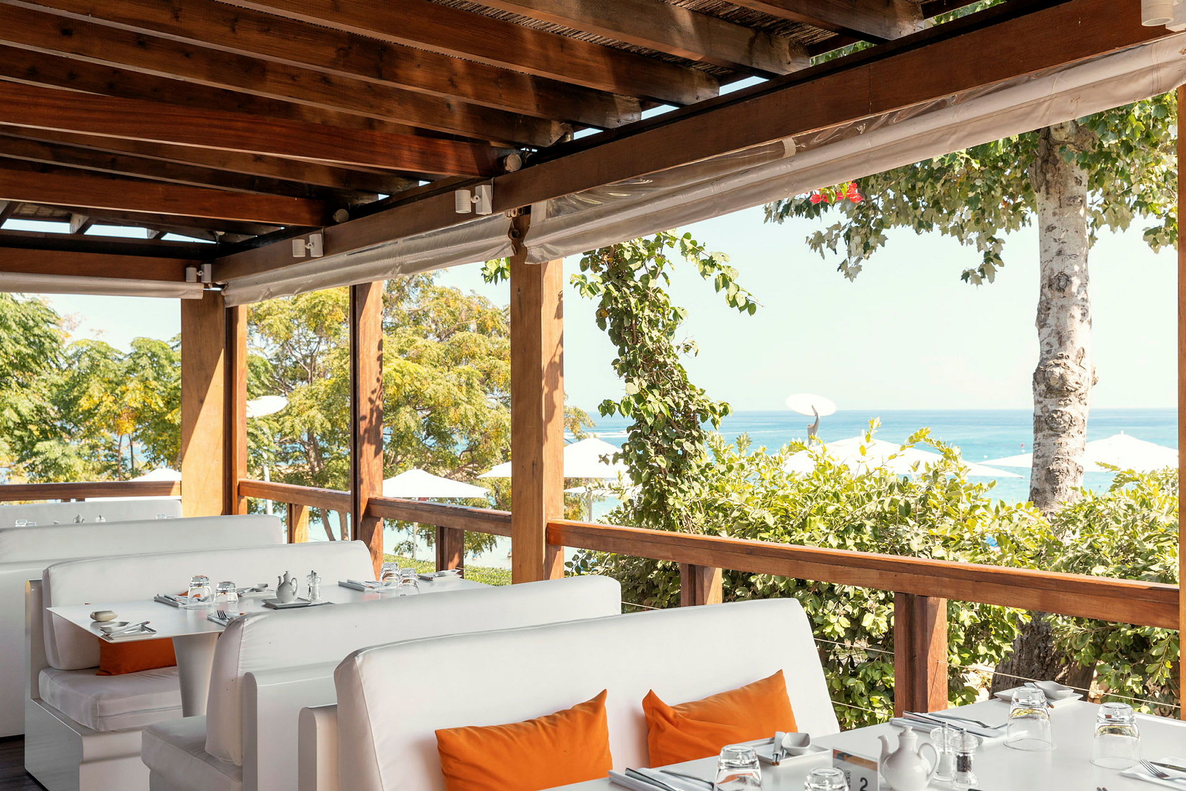 Bild på en restaurang med vita soffor och bord med utsikt över havet och grönska. (AI-genererad text)