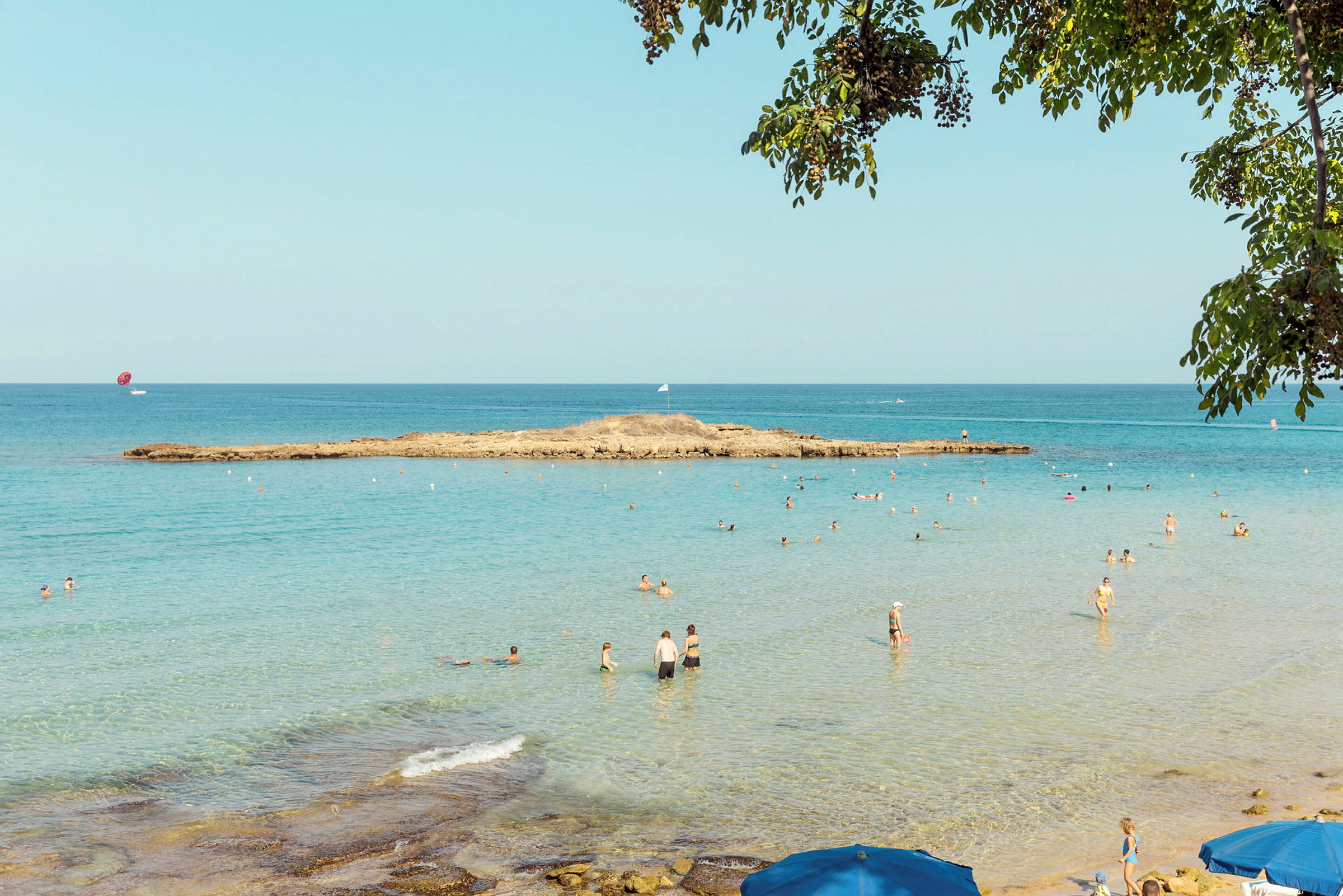 Strand med turkost vatten, där människor badar och småparasoll syns i förgrunden. (AI-genererad text)