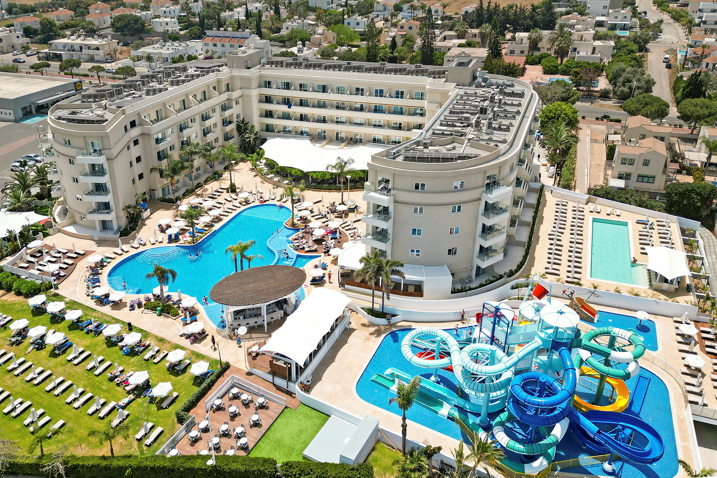 Ett hotellkomplex med flera pooler, solstolar och vattenrutschbanor, omgivet av grönområden och byggnader. (AI-genererad text)