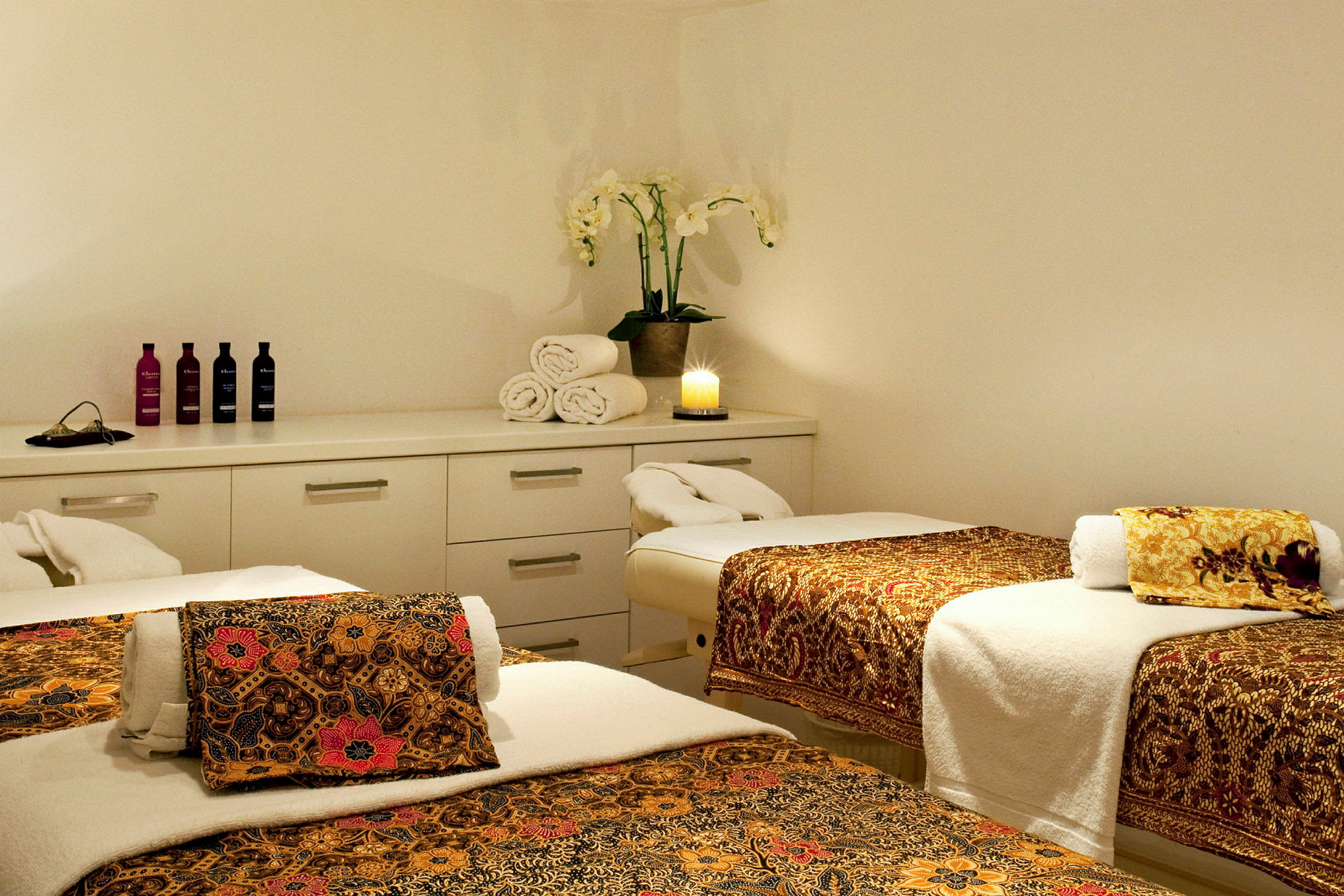 Ett spa-rum med tre massagebänkar klädda med tyg med orientaliska mönster, handdukar och en växt samt en tänd ljus på en byrå. (AI-genererad text)