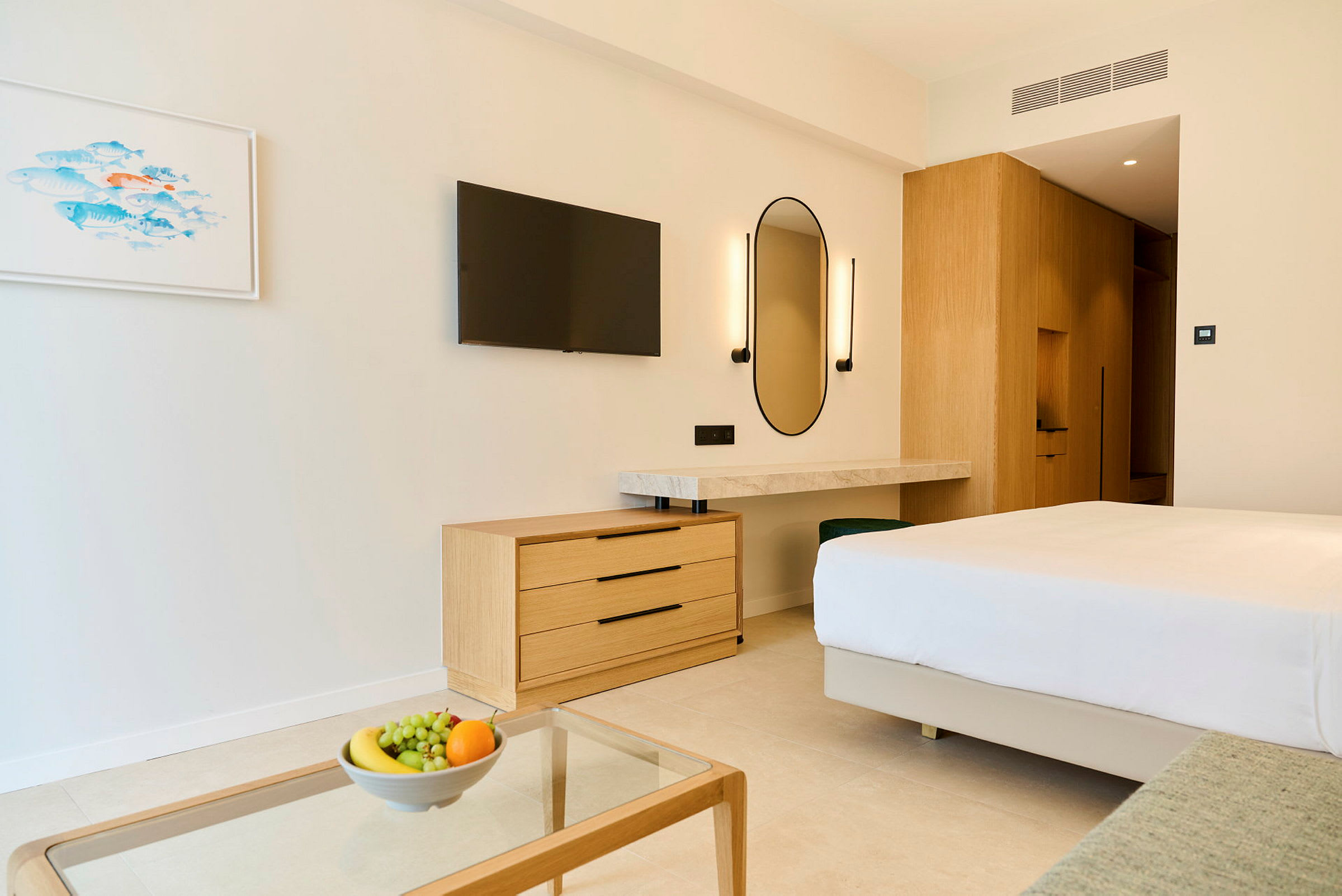 Ett modernt hotellrum med en dubbelsäng, en byrå, en väggmonterad TV och ett skrivbord med spegel. På ett glasbord står en skål med frukt. (AI-genererad text)