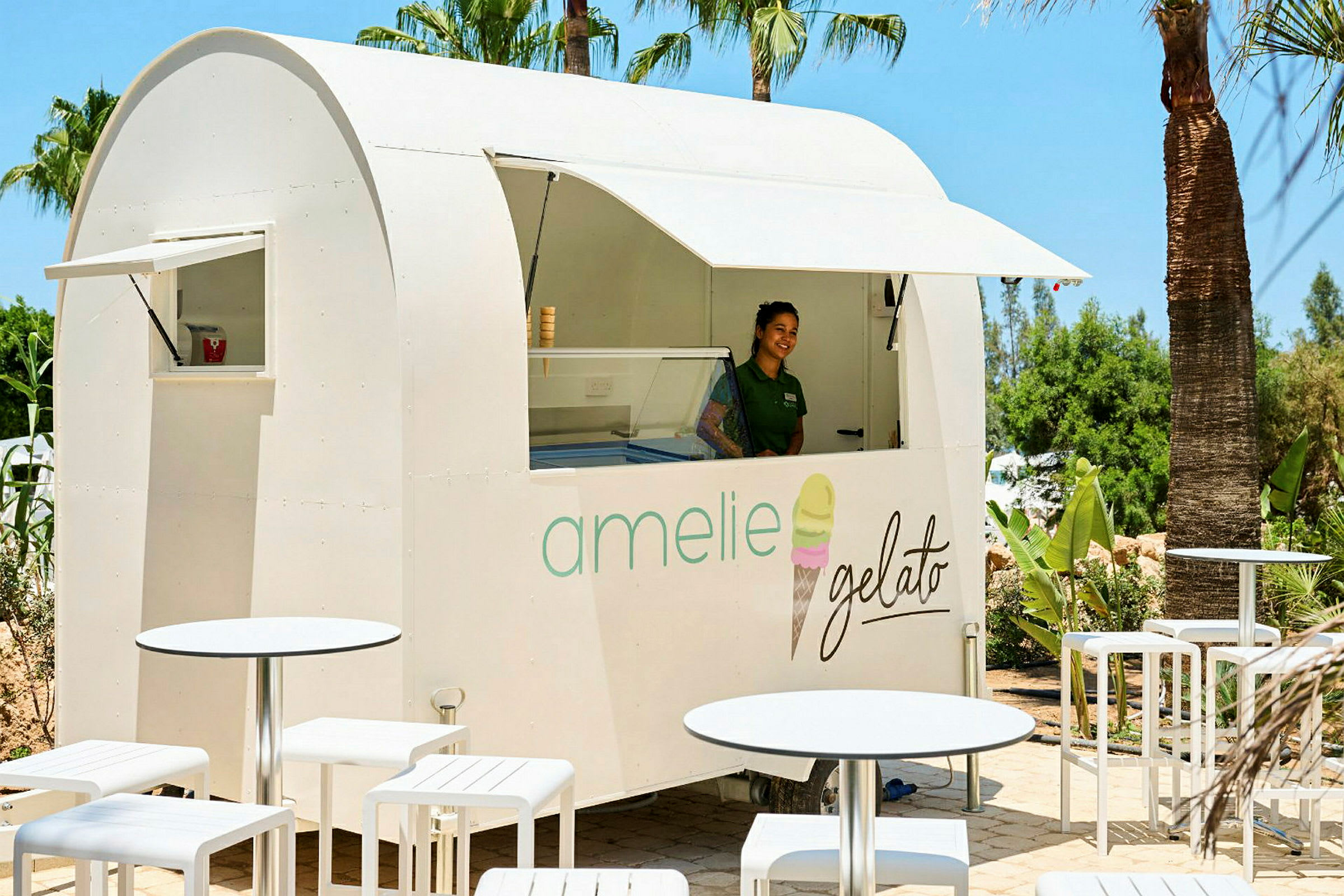 En vit glasskiosk med texten "amelie gelato" står bland palmer, omgiven av vita utemöbler. (AI-genererad text)