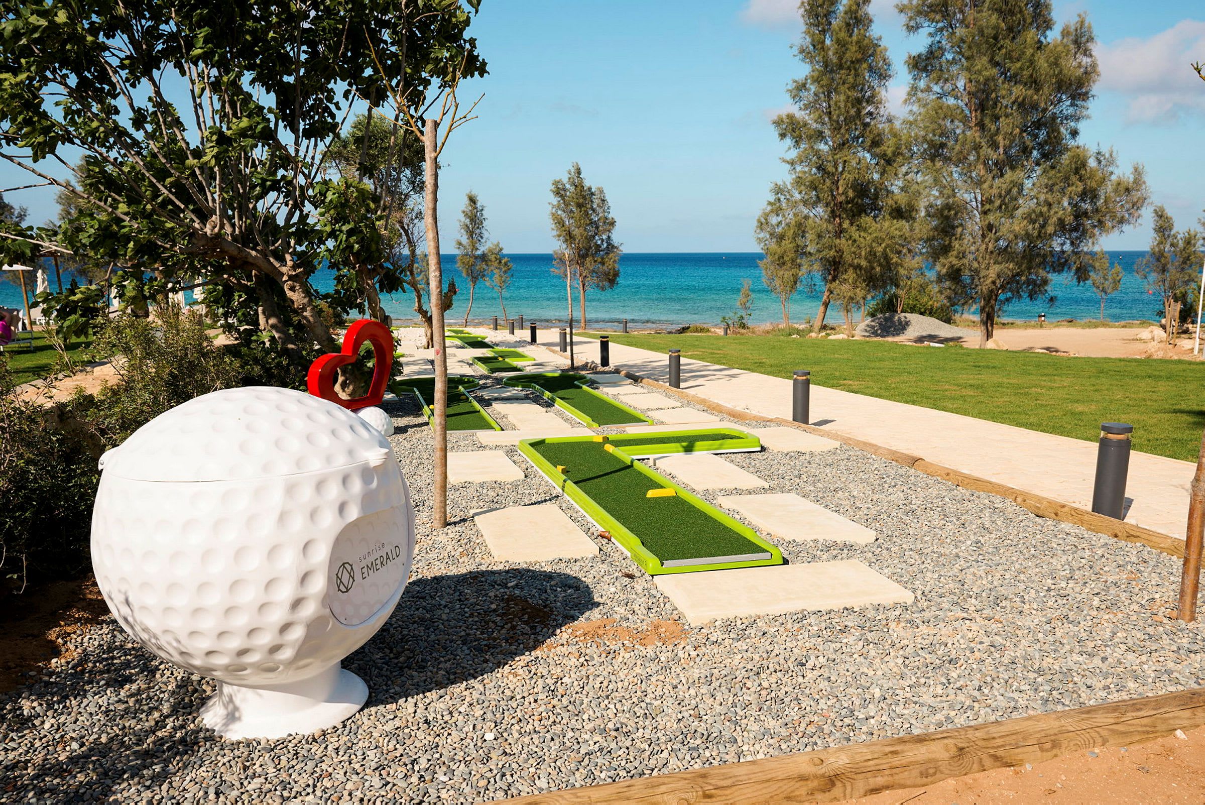 En minigolfbana med gröna banor ligger nära stranden med blått hav i bakgrunden. En stor golfbollsskulptur syns på sidan. (AI-genererad text)