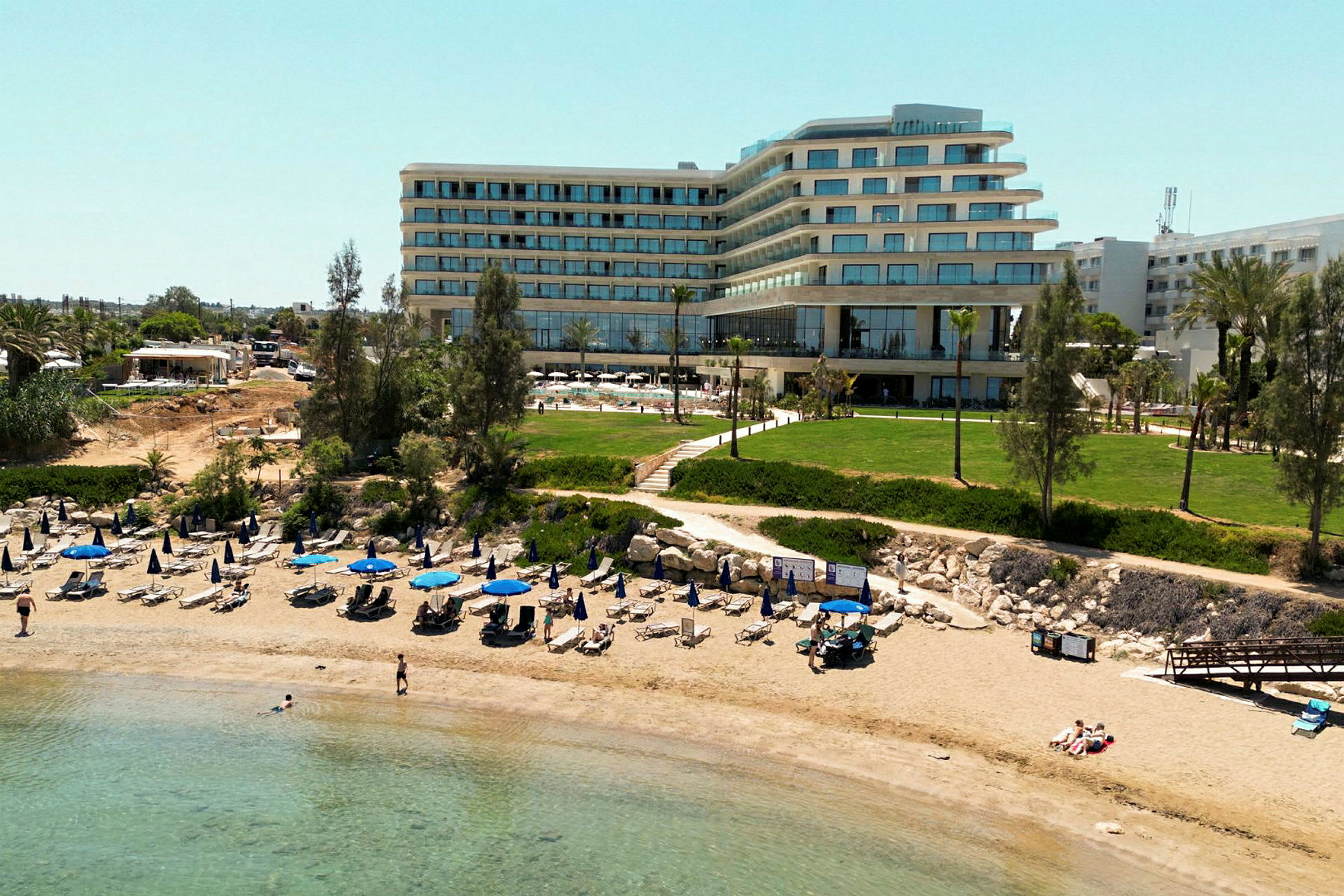 Bild av en sandstrand med uppställda parasoller och solstolar framför en modern hotellbyggnad och grönområden. (AI-genererad text)