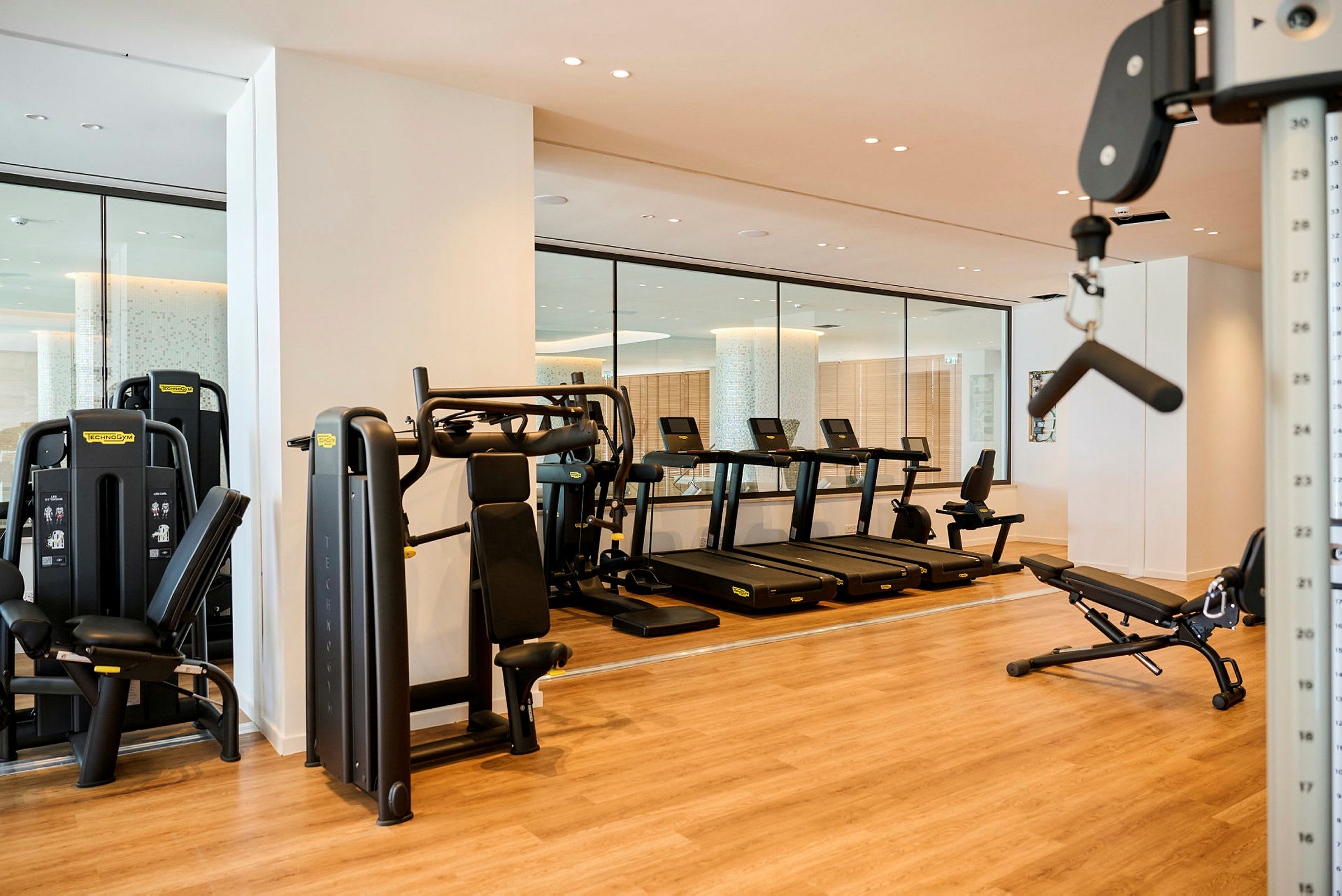 Ett modernt gym med träningsutrustning som löpband, styrketräningsmaskiner och bänkar på ett trägolv. (AI-genererad text)