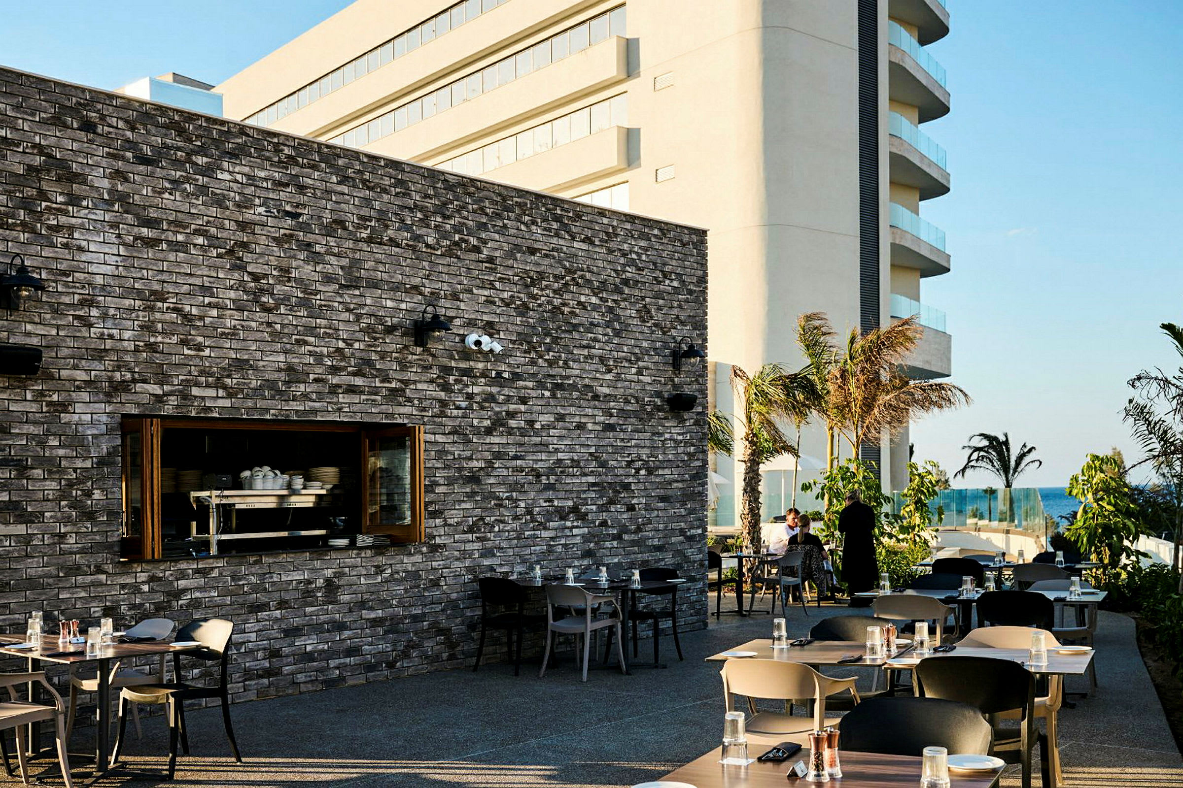 Utomhusterrass vid en restaurang med bord och stolar. En stor byggnad syns i bakgrunden, med palmer och havet i avstånd. (AI-genererad text)