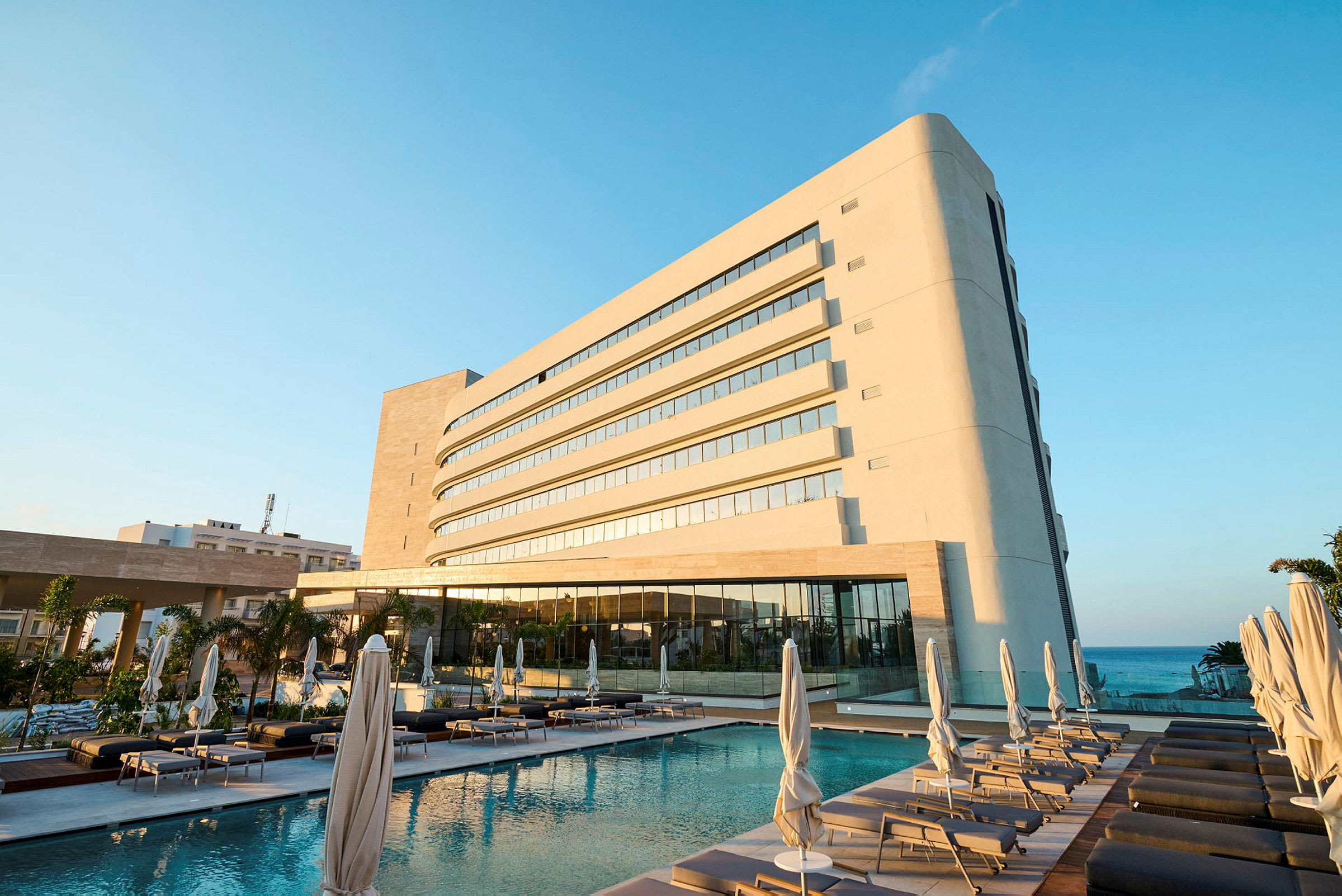 En modern hotellbyggnad bredvid en pool med solstolar och parasoller. Havet syns i bakgrunden. (AI-genererad text)