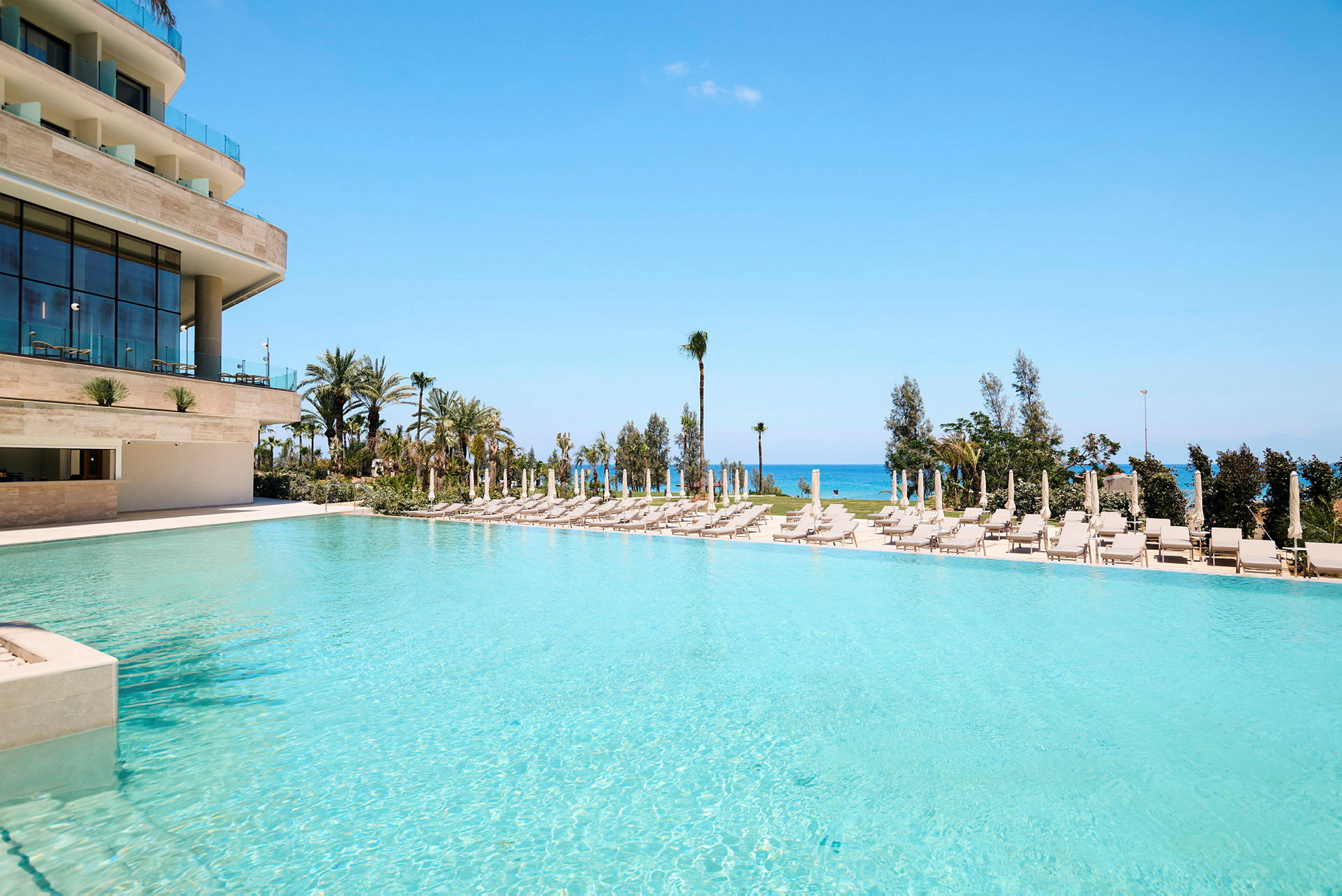 En swimmingpool vid en modern byggnad med solstolar och parasoller runtom, samt havet i bakgrunden. (AI-genererad text)