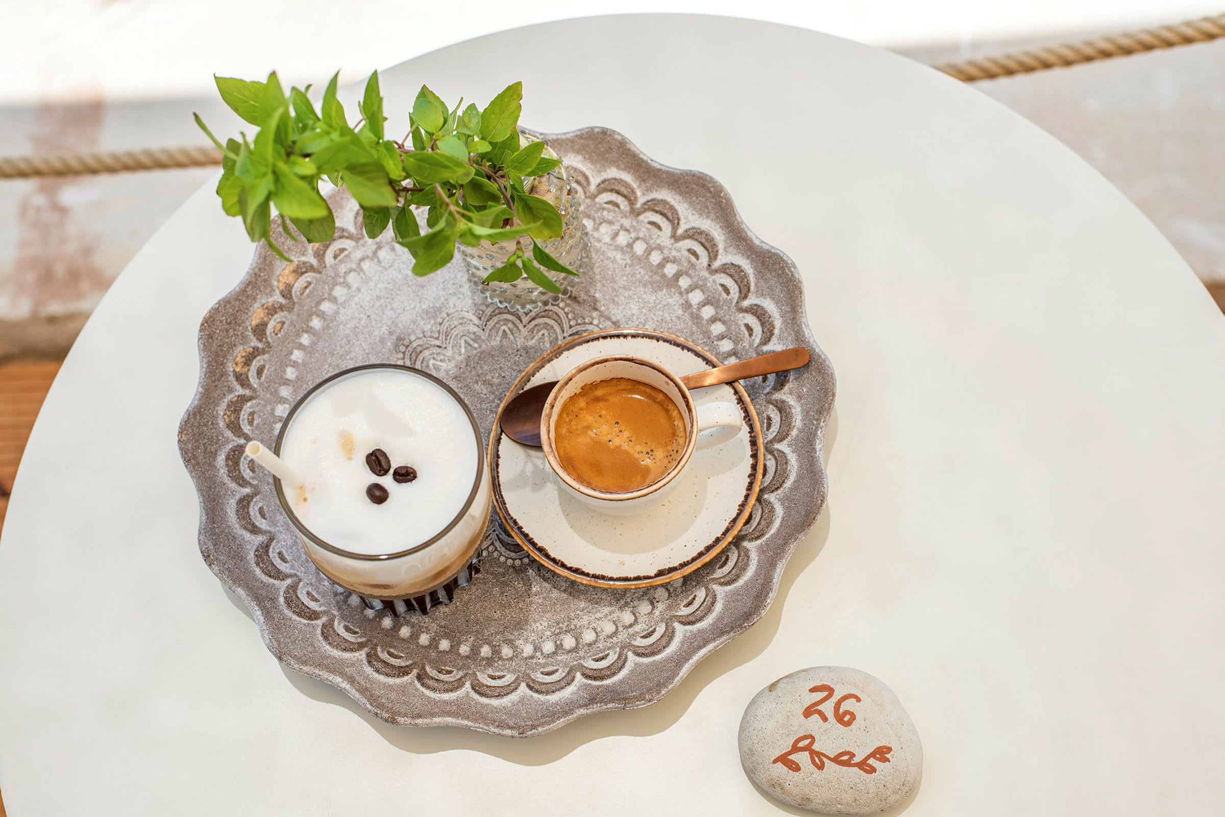 En detaljbild på en bricka med en kopp kaffe, en milkshake med kaffe, och en liten glasvas med gröna blad på ett bord. (AI-genererad text)