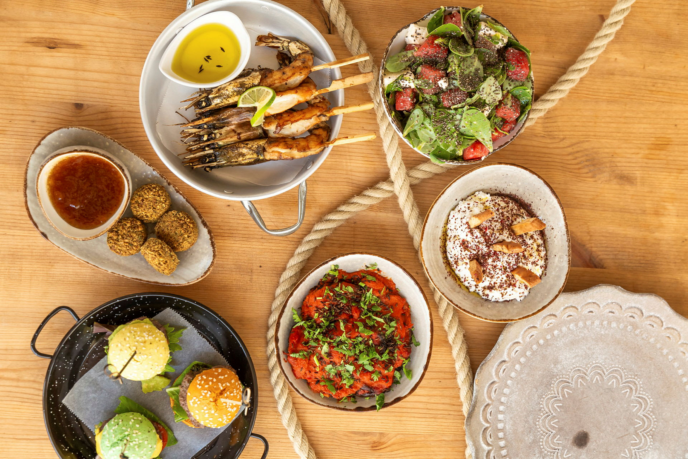 En mängd olika maträtter såsom räkor på spett, sallad, falafel, burgare, samt dippsåser och tillbehör på ett träbord. (AI-genererad text)