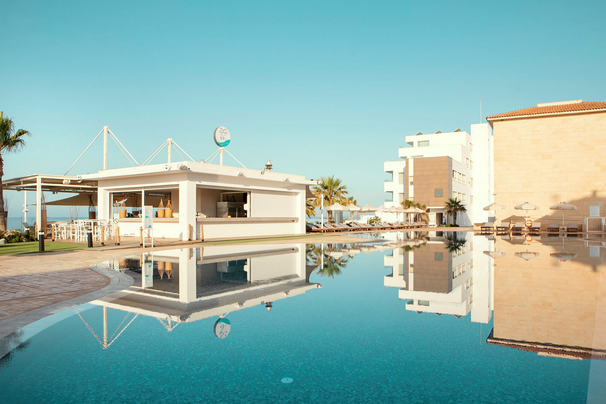 En pool med klarblått vatten omgiven av solstolar och byggnader under en blå himmel. (AI-genererad text)