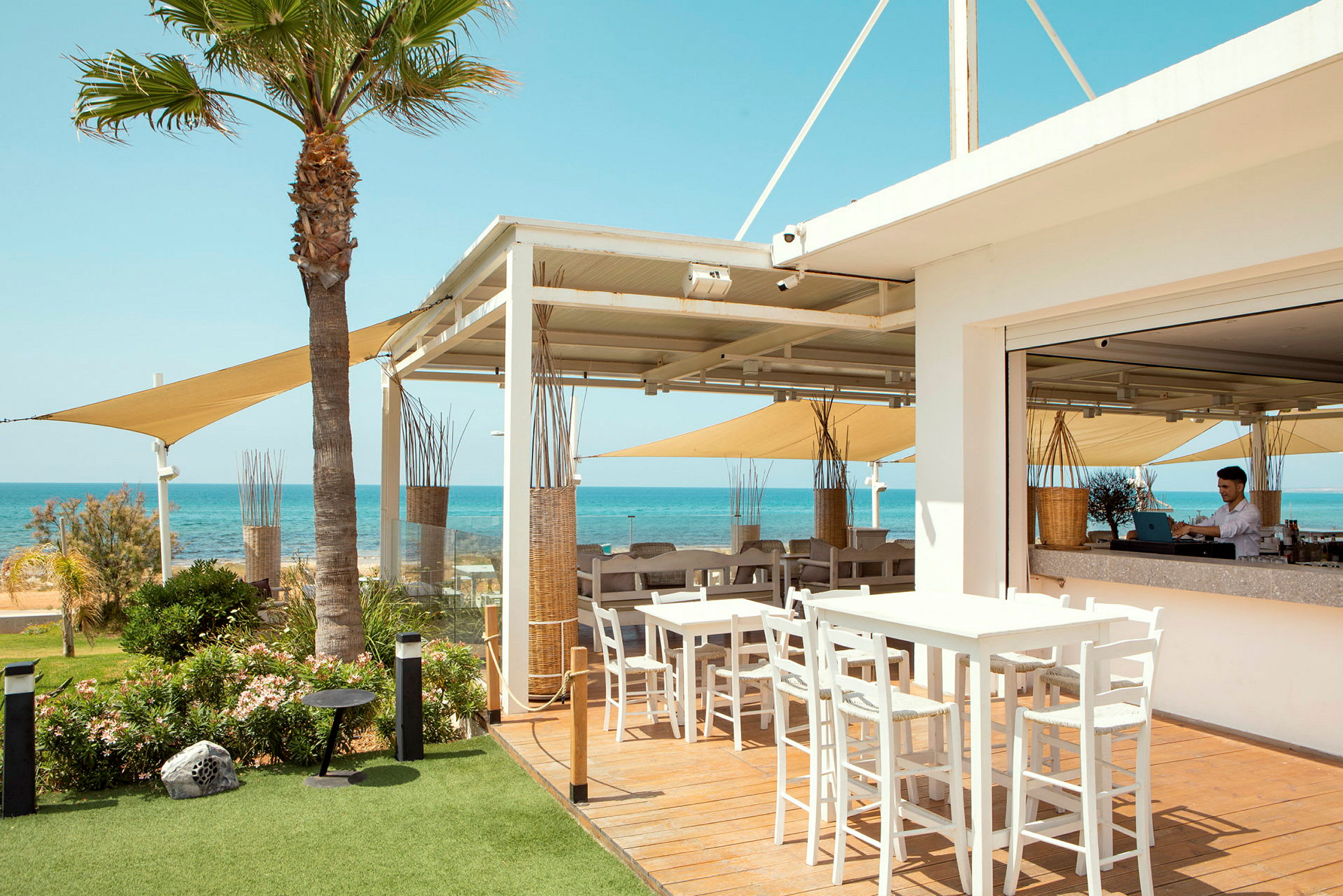 En utomhusrestaurang med bord och stolar, utsikt över havet och klarblå himmel. (AI-genererad text)