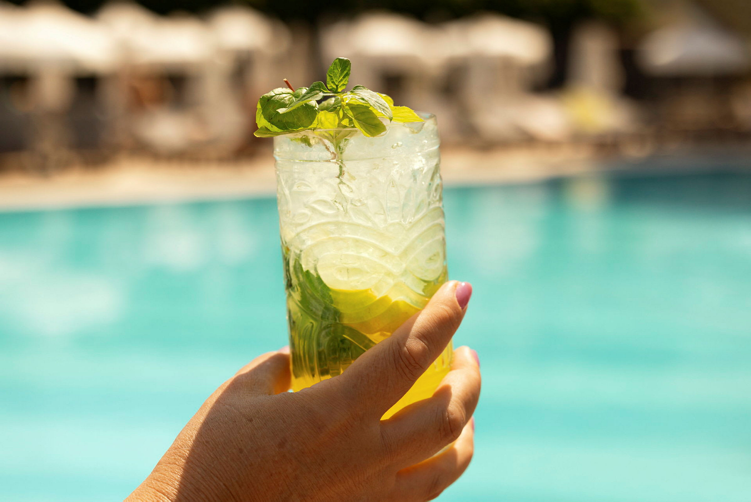En hand håller ett glas med en kall dryck, dekorerad med mynta och citron, framför en utomhuspool. (AI-genererad text)