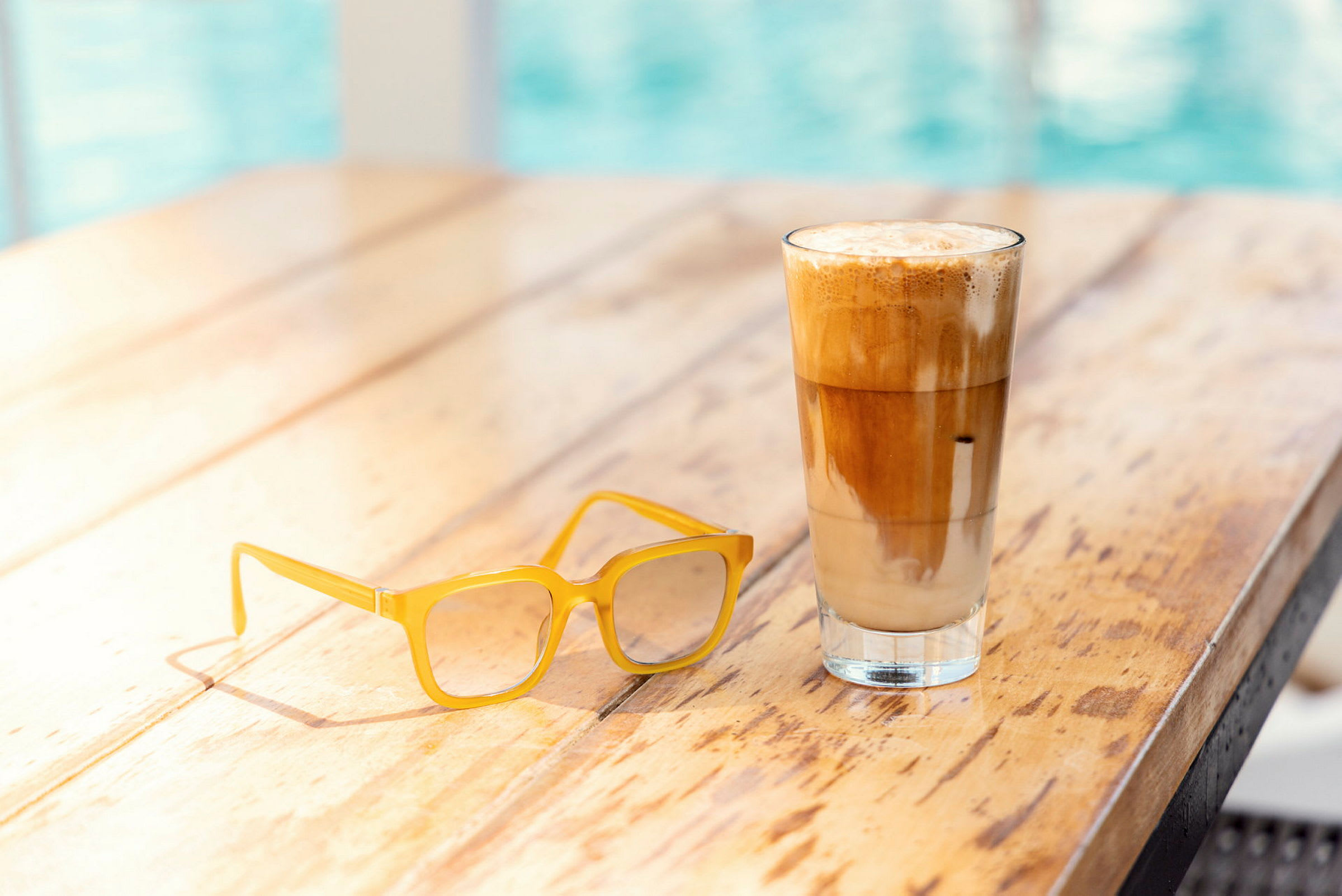 Ett träbord med ett par gula glasögon och ett glas iskaffe eller latte. (AI-genererad text)