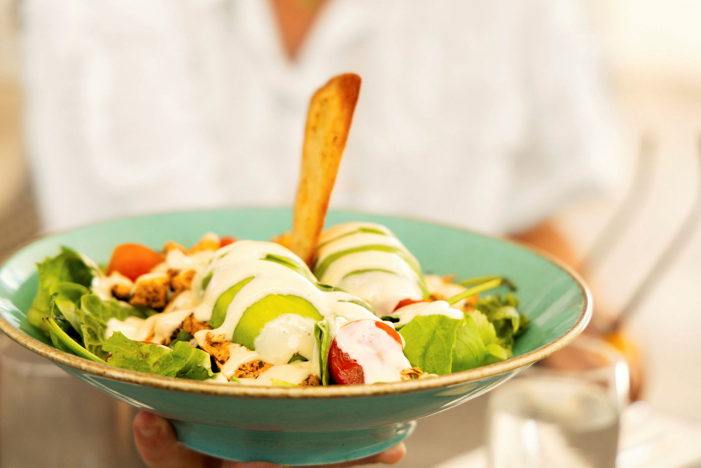 En sallad serverad i en turkos skål, innehållande grönsaker, avokado, dressing och en brödpinne. (AI-genererad text)