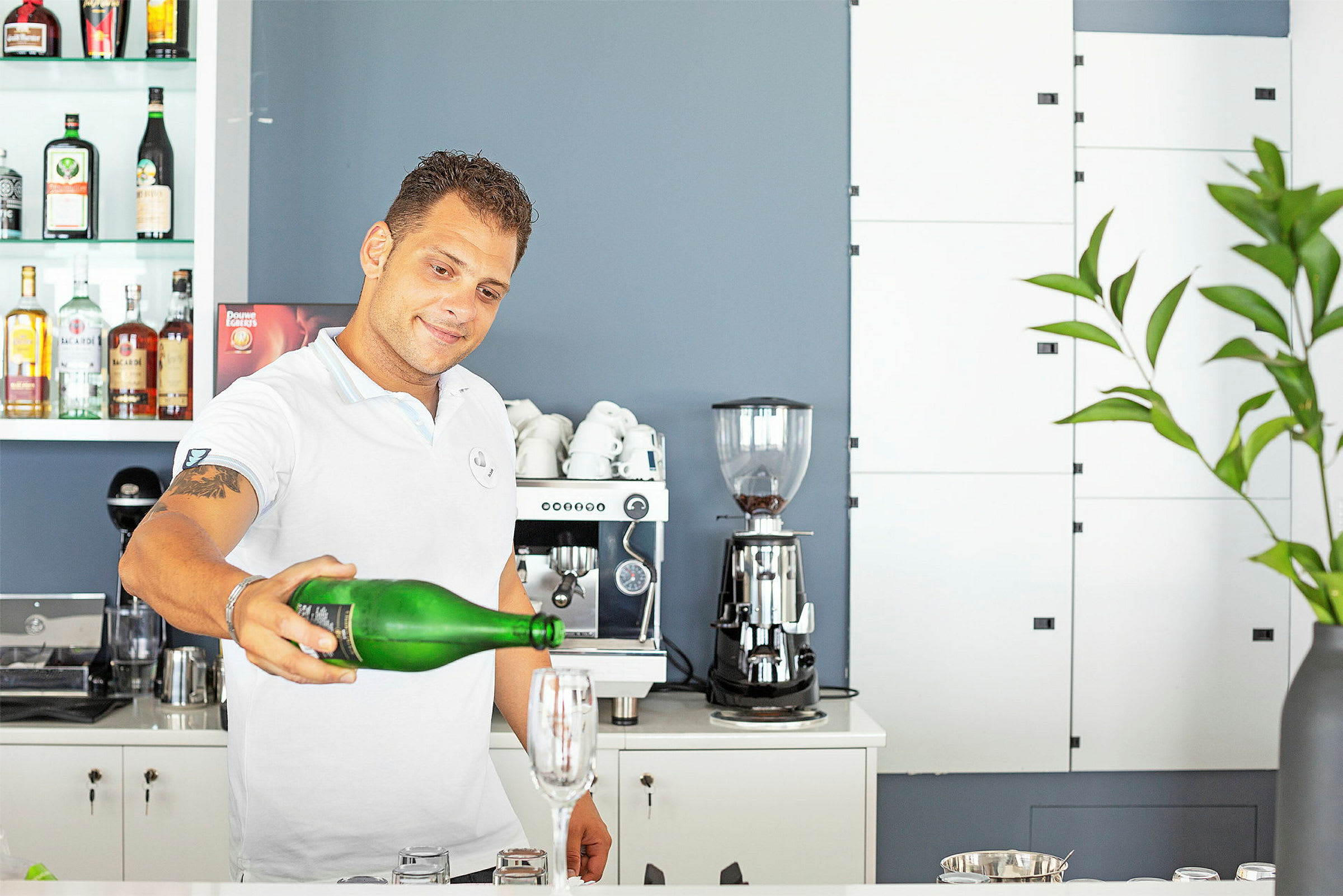 En person håller en grön flaska och häller vätska i ett glas på en bardisk, omgivande av en kaffemaskin och flaskor. (AI-genererad text)