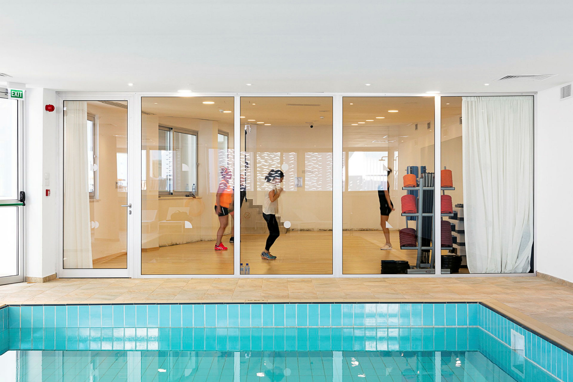 Glasdörrar till ett gym med personer som tränar, sett från ett rum med en pool. (AI-genererad text)